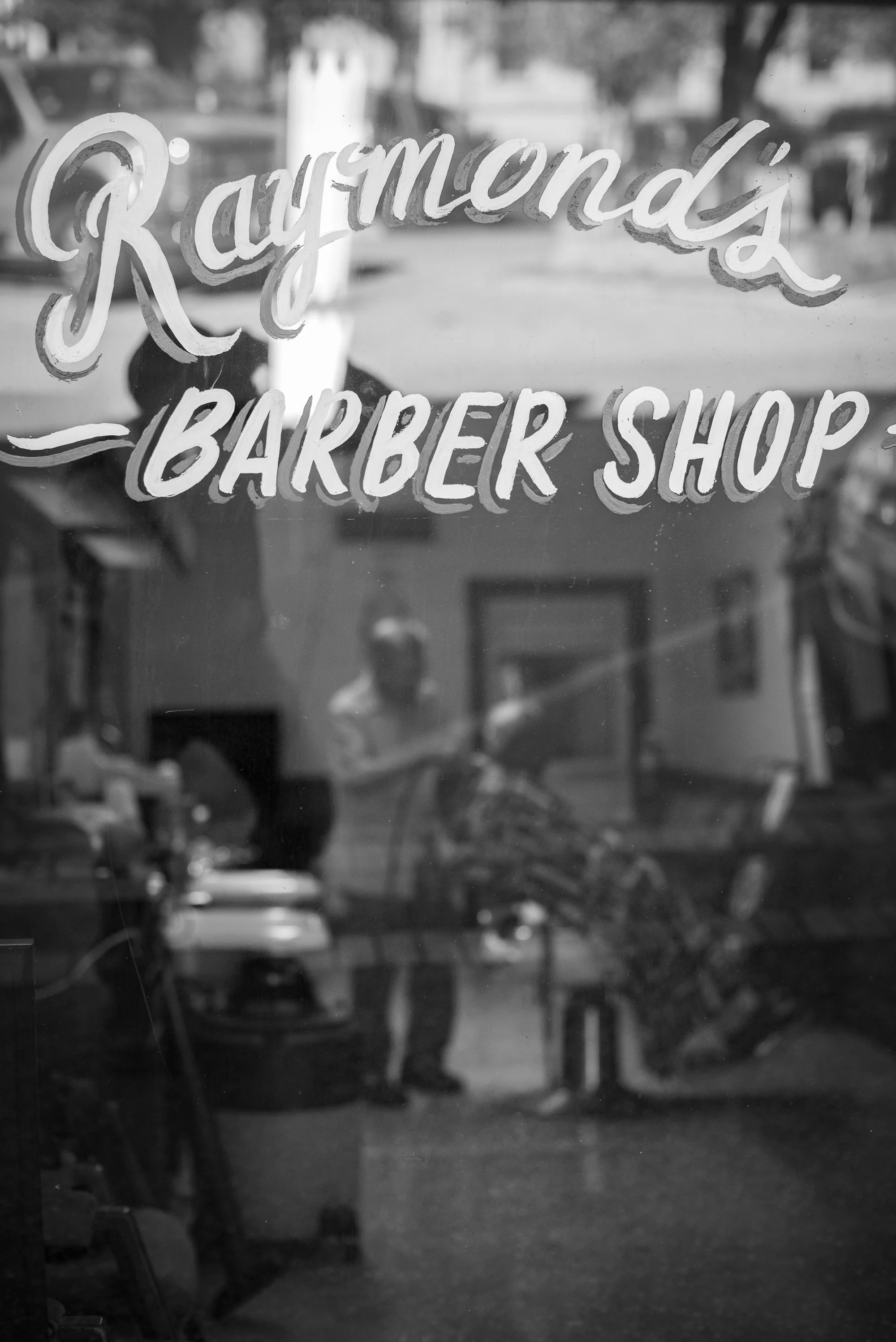 Hand-painted “Raymond’s Barber Shop” lettering on the front window in Lockhart, Texas, with reflections of the barbershop interior.