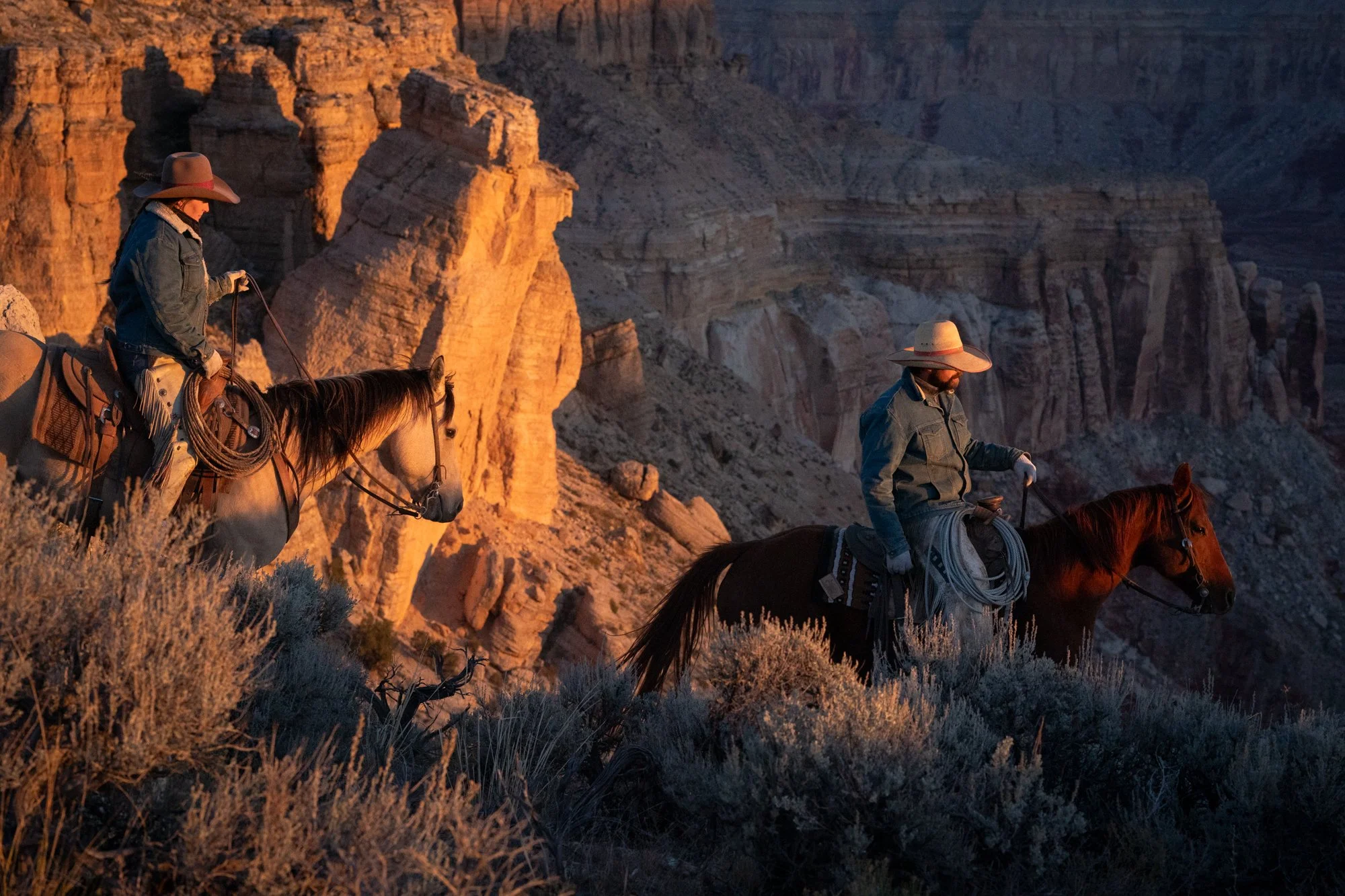 Two working cowboys riding horseback through canyon terrain at Diamond A Ranch in Arizona during golden hour light