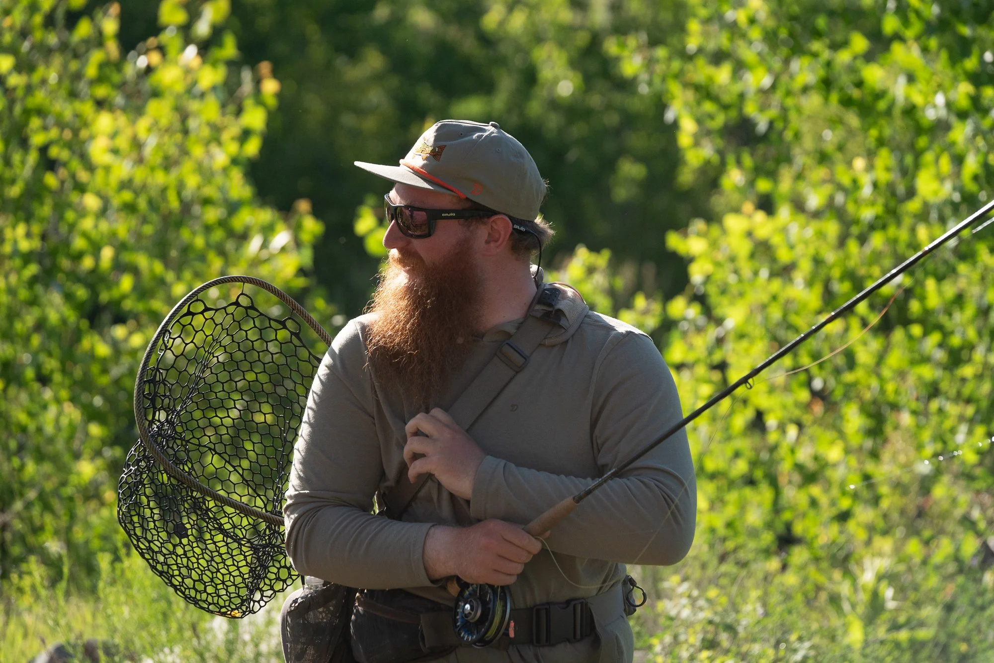 A fly fishing angler holds a rod and landing net while standing along a green riverbank