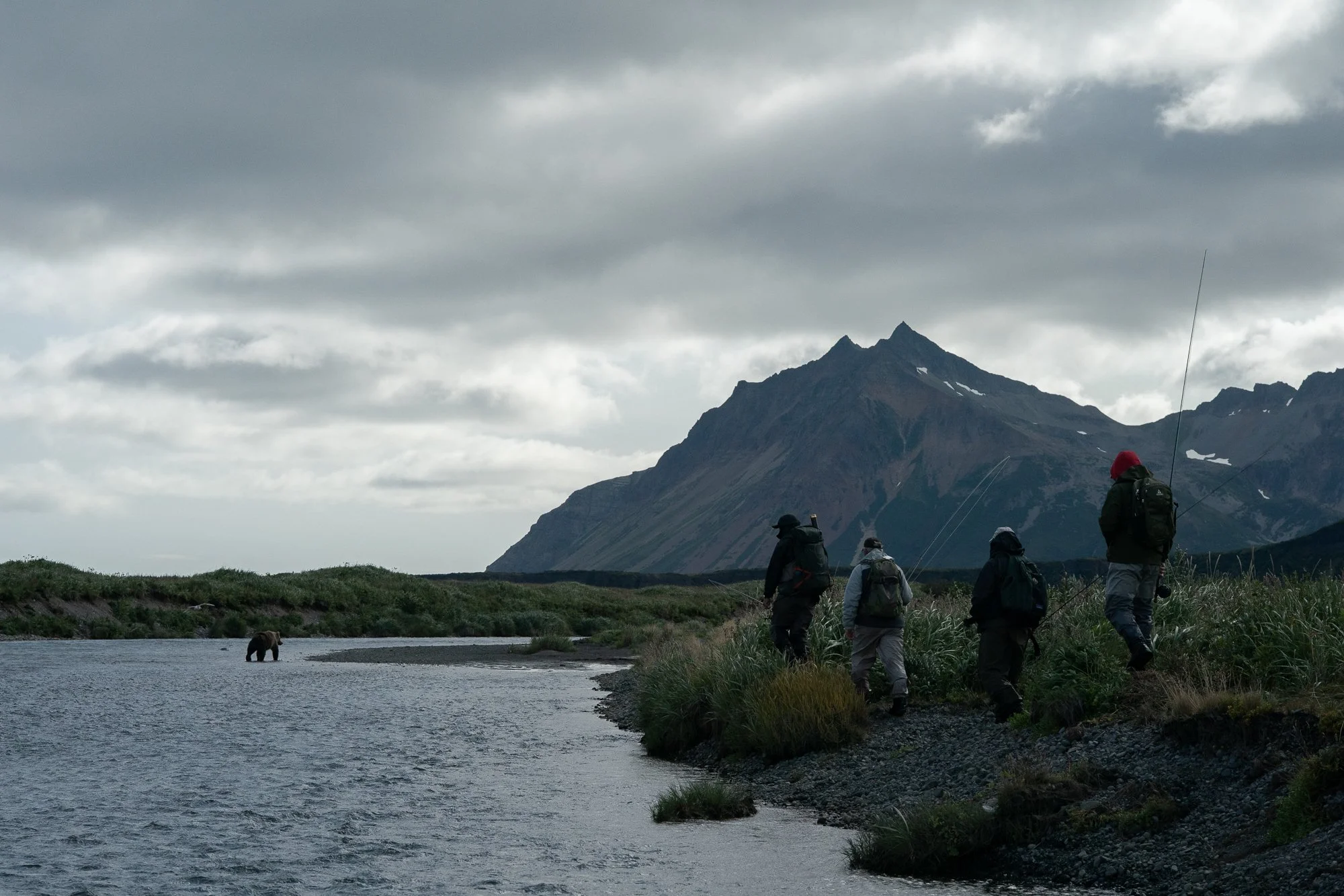 Fly fishermen walking along a river with a grizzly bear nearby and mountains behind them