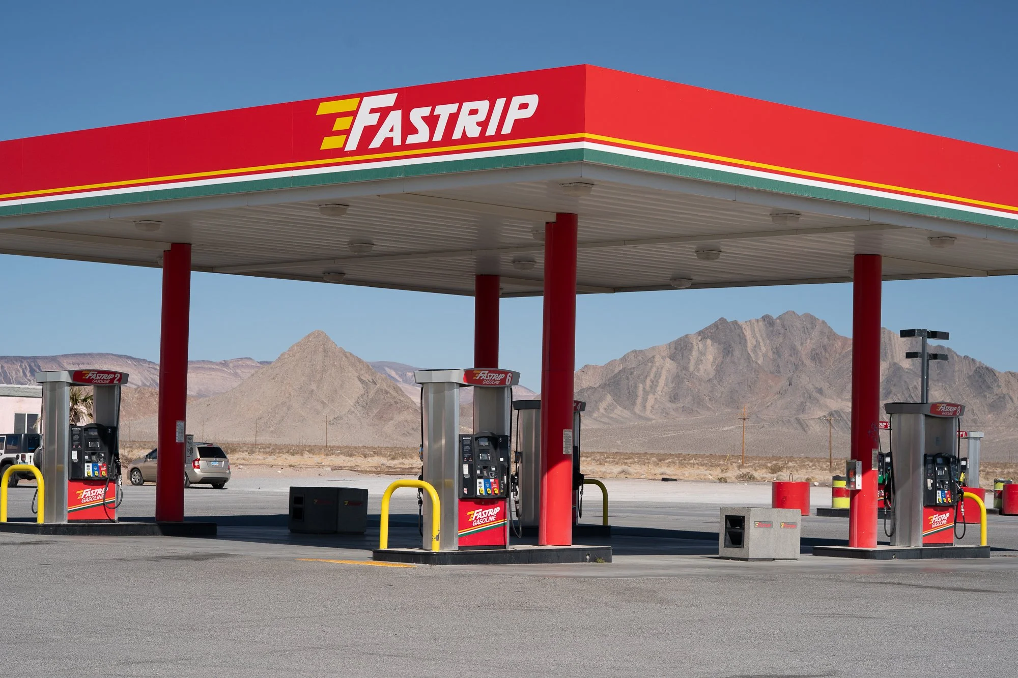 Empty Fastrip gas station canopy with fuel pumps and no customers, photographed against a clear blue sky and a large rocky Nevada desert mountain range.