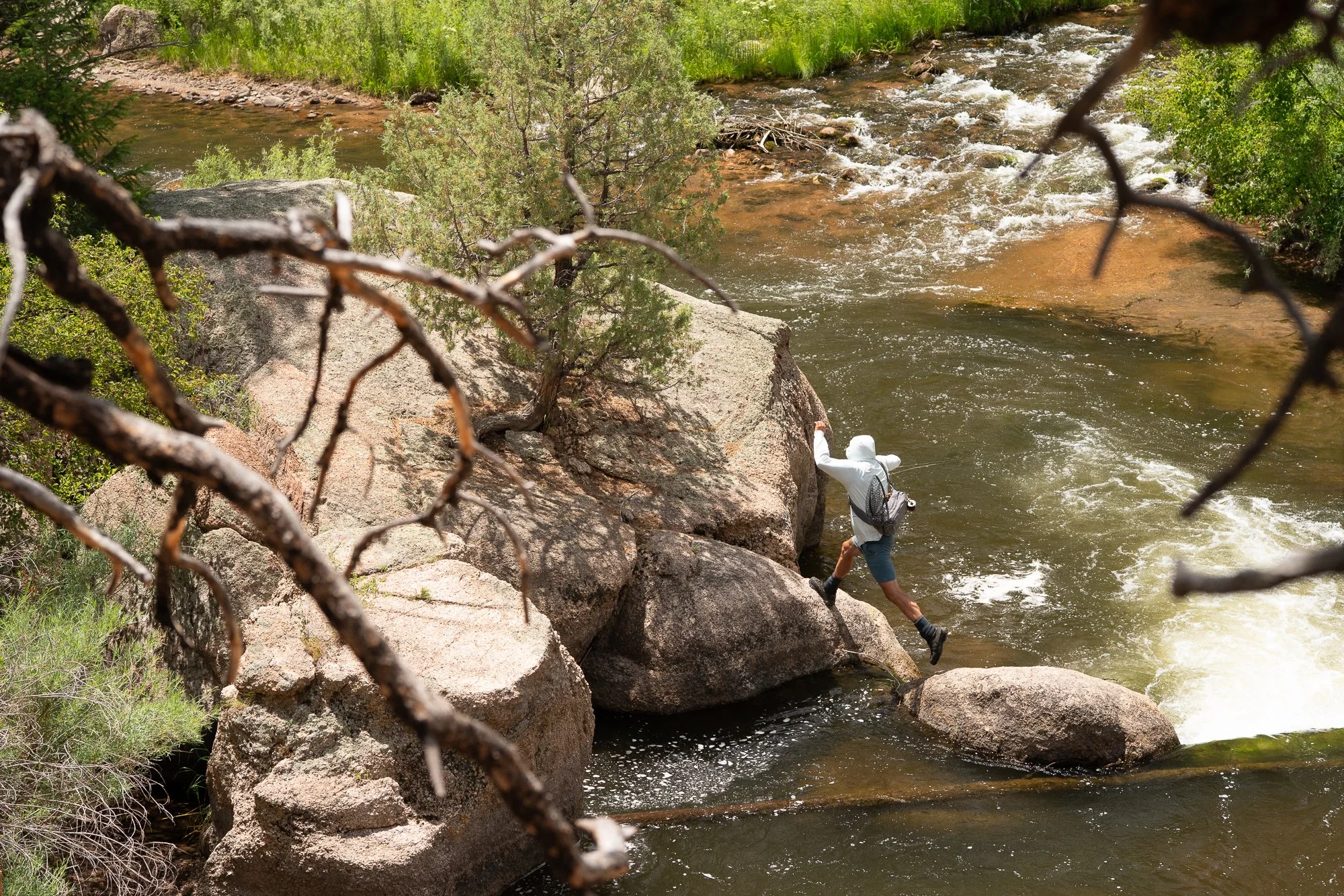 A fly angler steps across rocks beside moving river water with a rod in hand