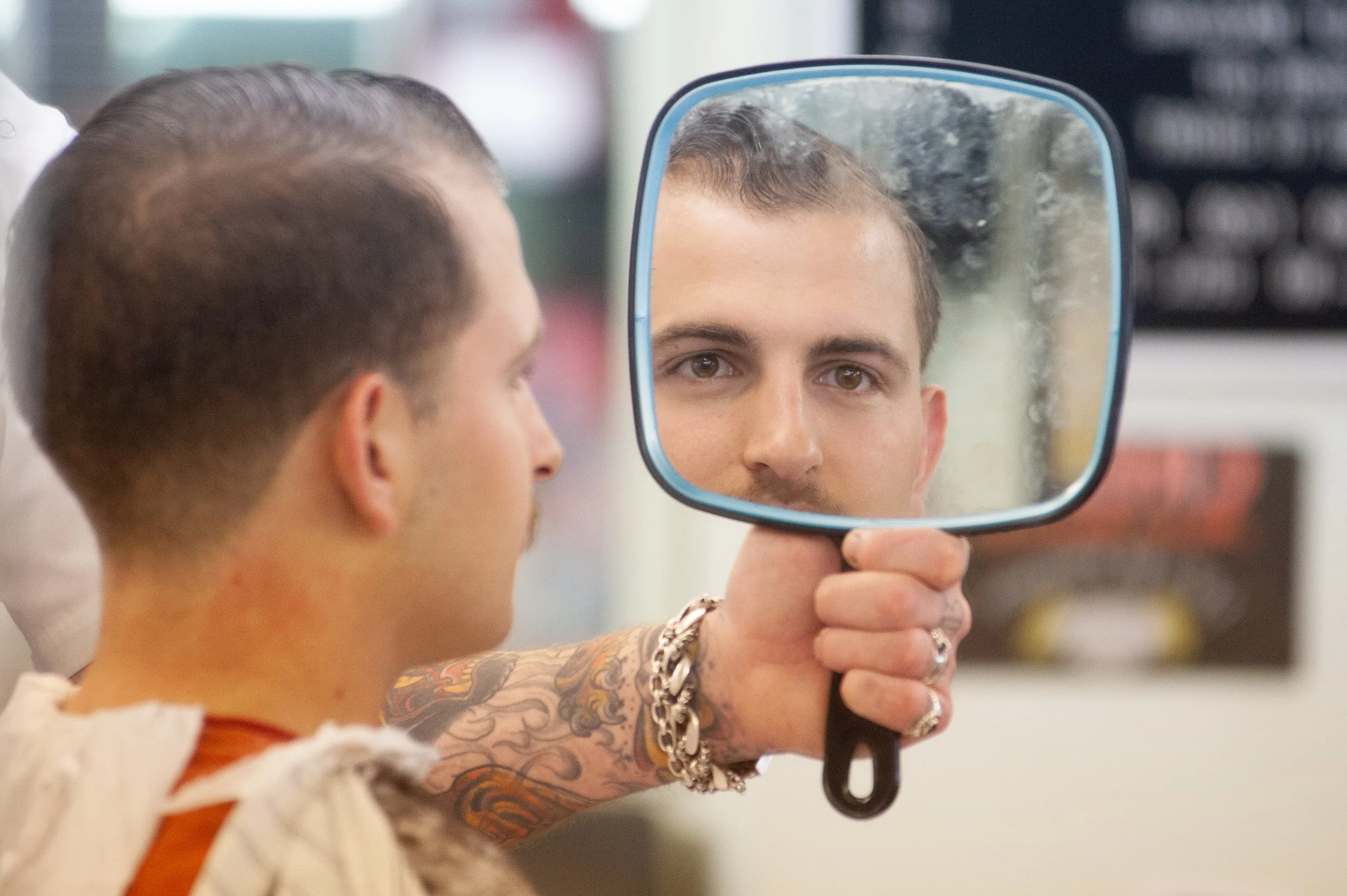 Tattooed barber's hand holding a square hand mirror up to a caped client checking his fresh haircut at Sweeney Todd's Barbershop in Los Angeles