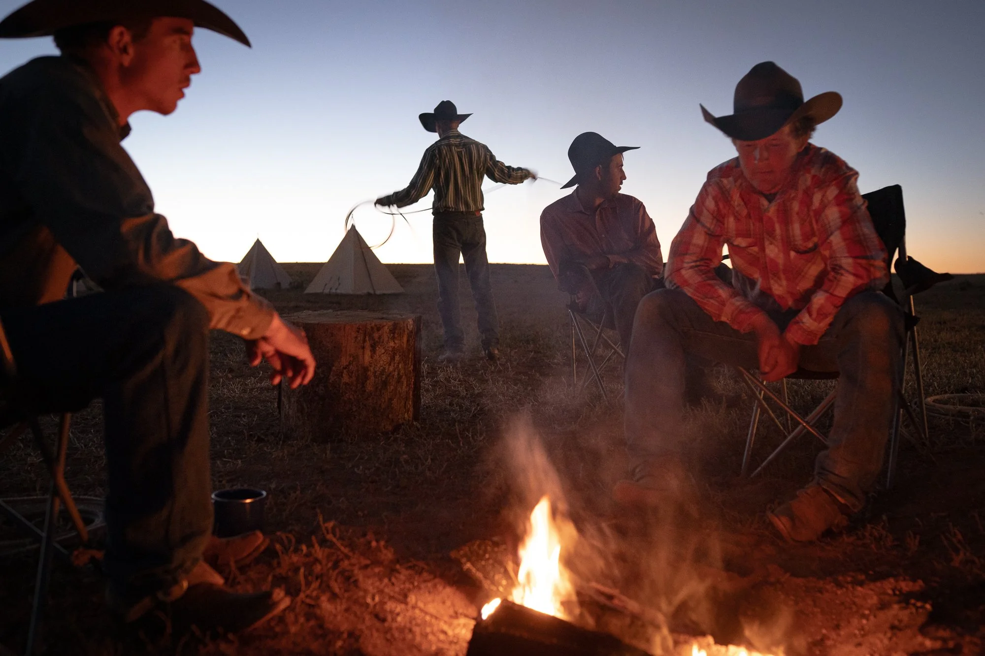 Cowboys gathered around campfire at night with tents and open landscape in background