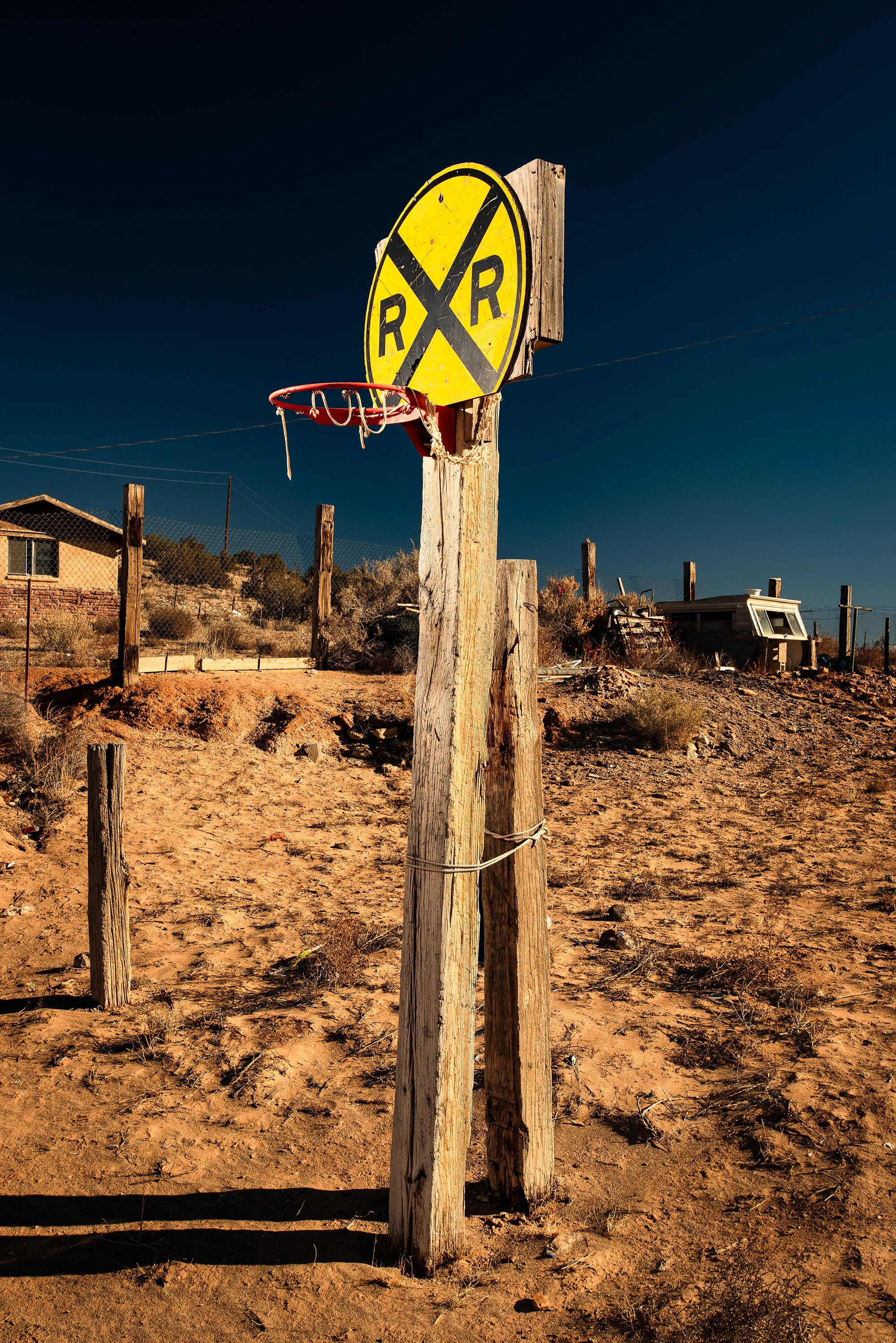 Basketball hoop mounted to a weathered wooden post beneath a yellow railroad crossing sign in a dry desert landscape.