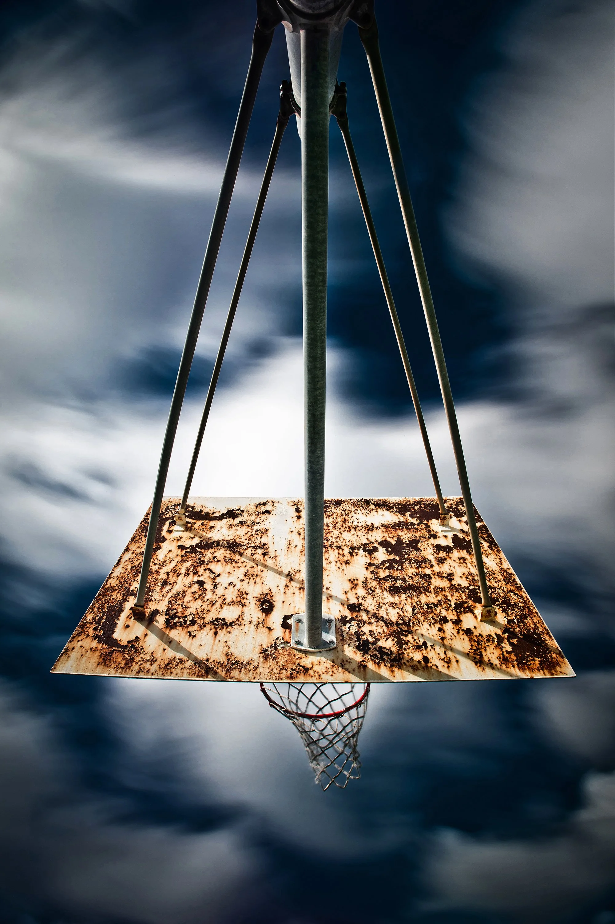 Rusted basketball backboard and hoop photographed from below against a cloudy sky.