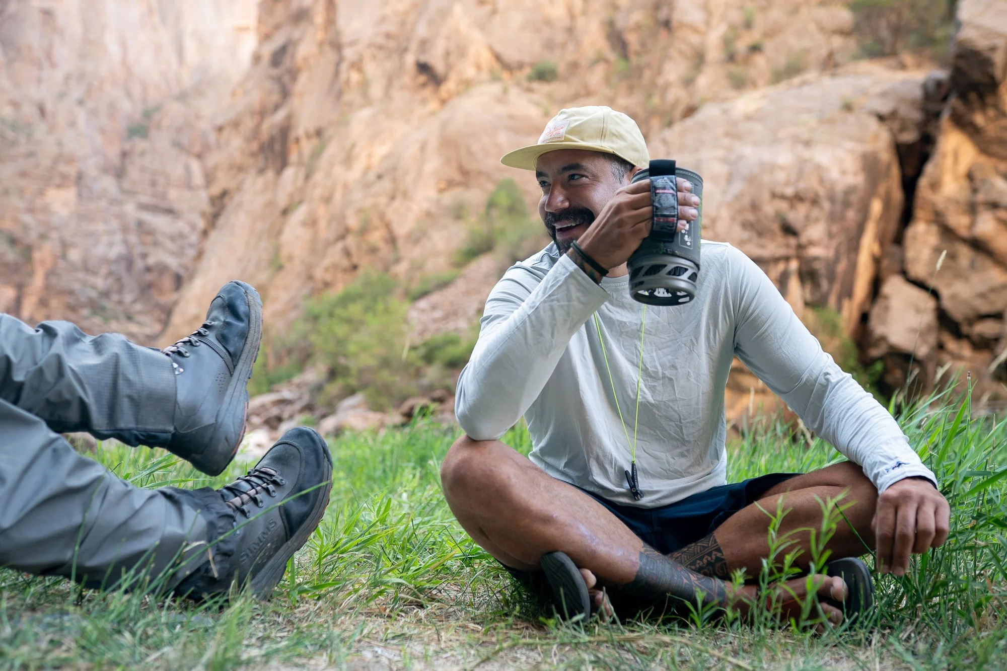 A man sits on the grass in Black Canyon holding a camp stove mug near the river