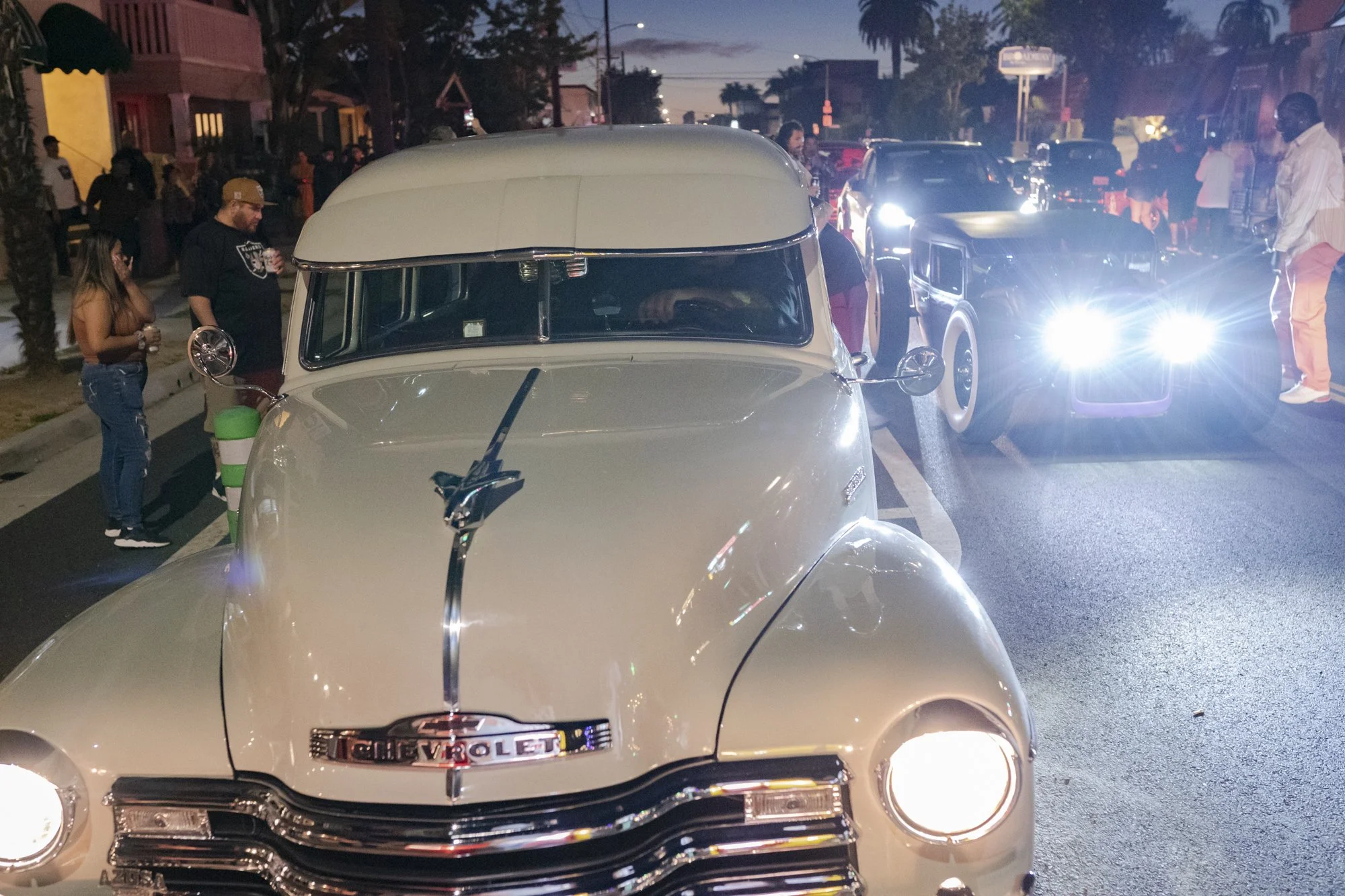 Cream-colored vintage Chevrolet truck facing the camera leading a procession of classic cars down a Long Beach street at night during the barbershop anniversary