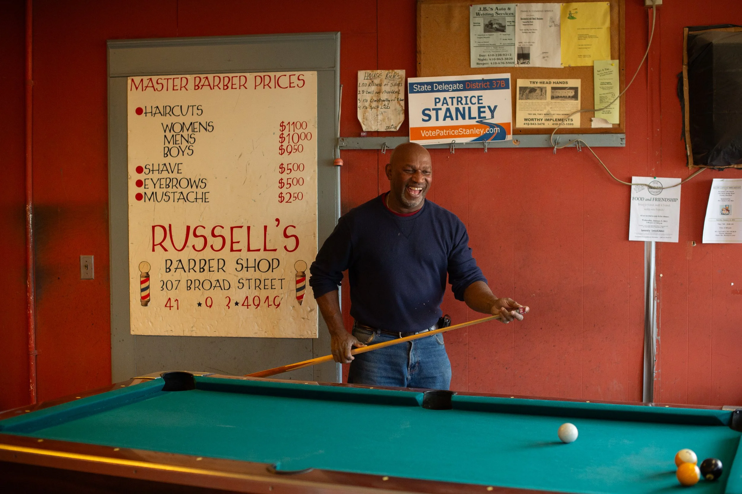 Man playing pool inside Russell’s Barbershop with price list and wall signs in the background