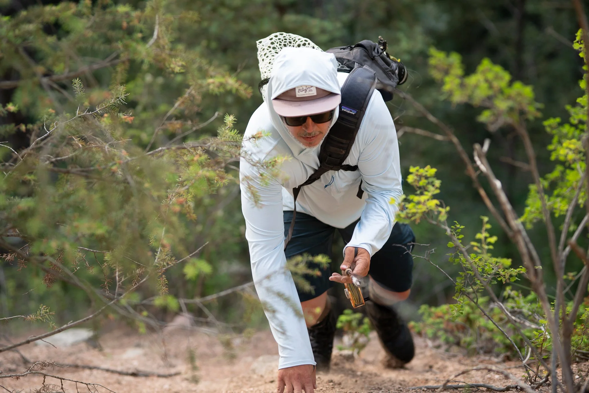 A fly angler moves through brush with a rod, pack, and landing net
