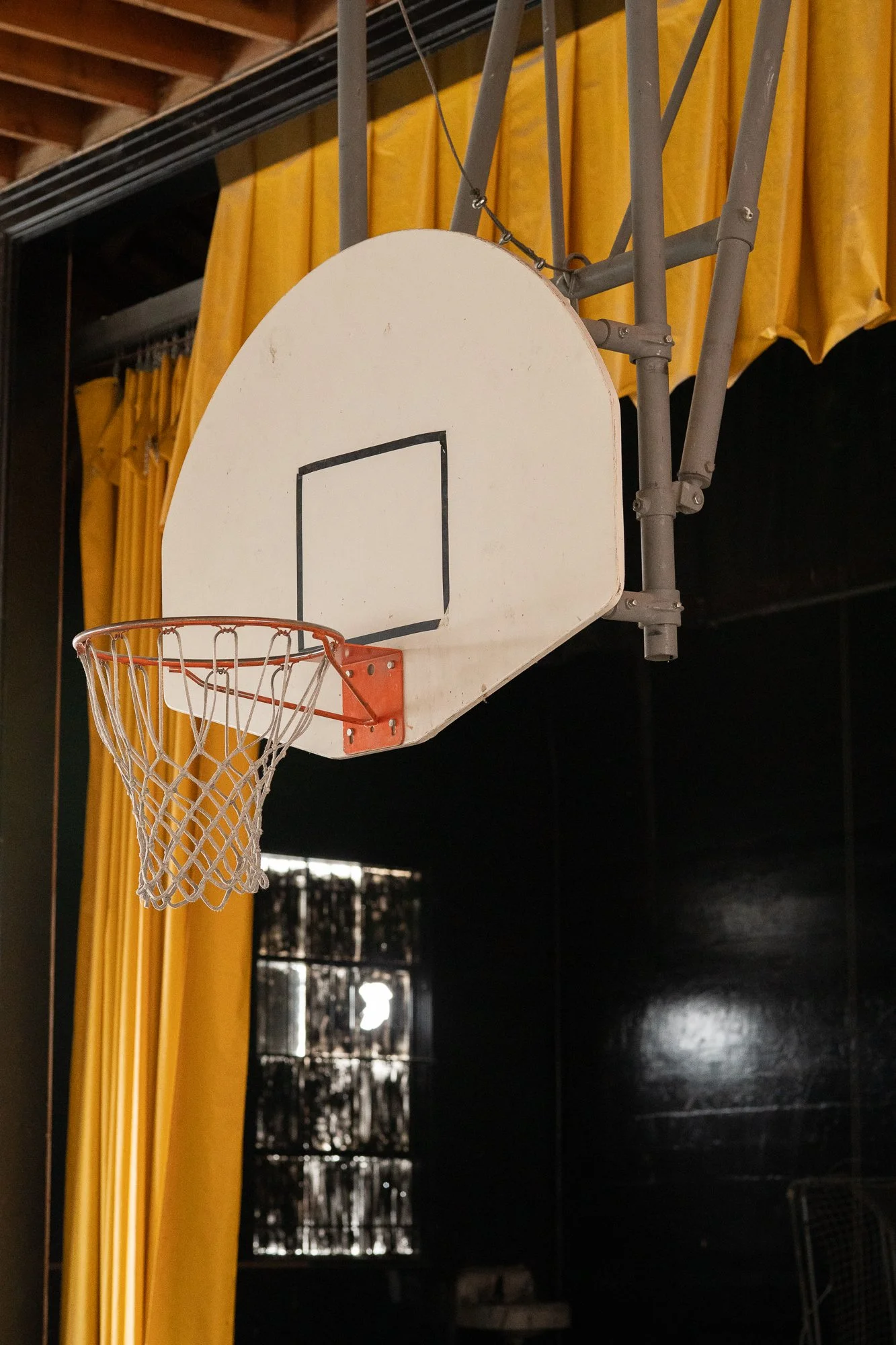Old basketball hoop and backboard mounted in a dim Nevada small town gymnasium, with yellow curtains and dark painted block walls visible in the background.