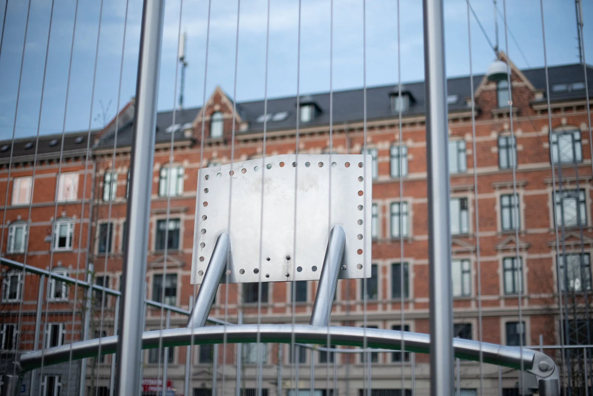 Outdoor basketball backboard photographed behind protective fencing on a European urban court.