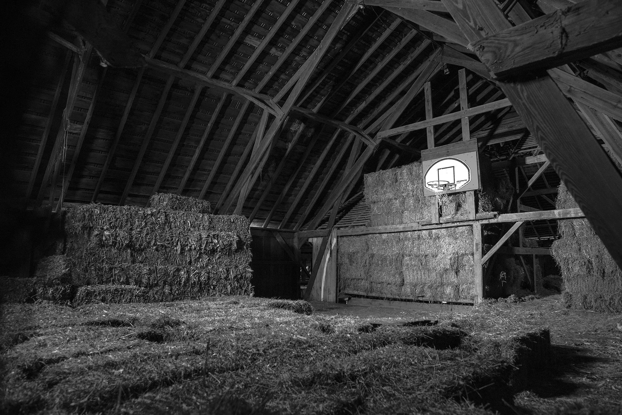 Basketball hoop hanging inside a wooden barn surrounded by stacked hay bales.