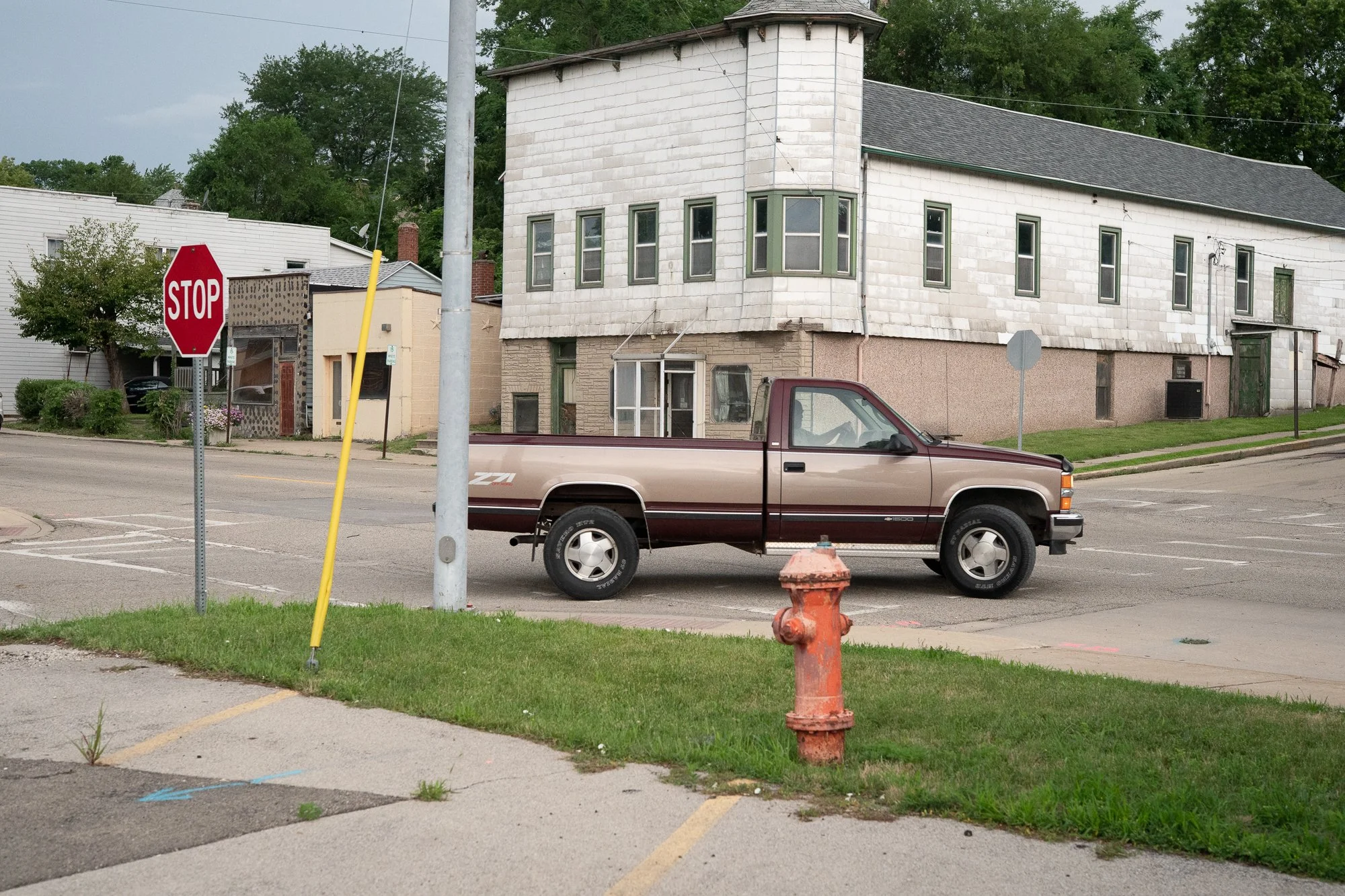 A maroon and tan Chevrolet pickup truck moves through a small-town intersection with older commercial buildings in the background.