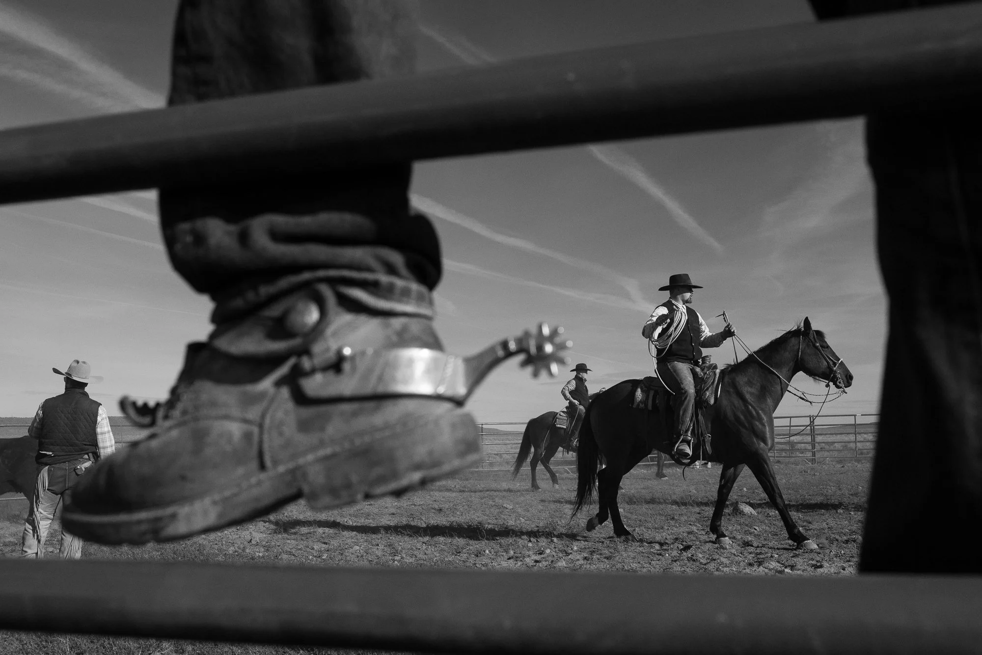 A black and white view of a cowboy boot and spur in the foreground while riders work nearby on horseback