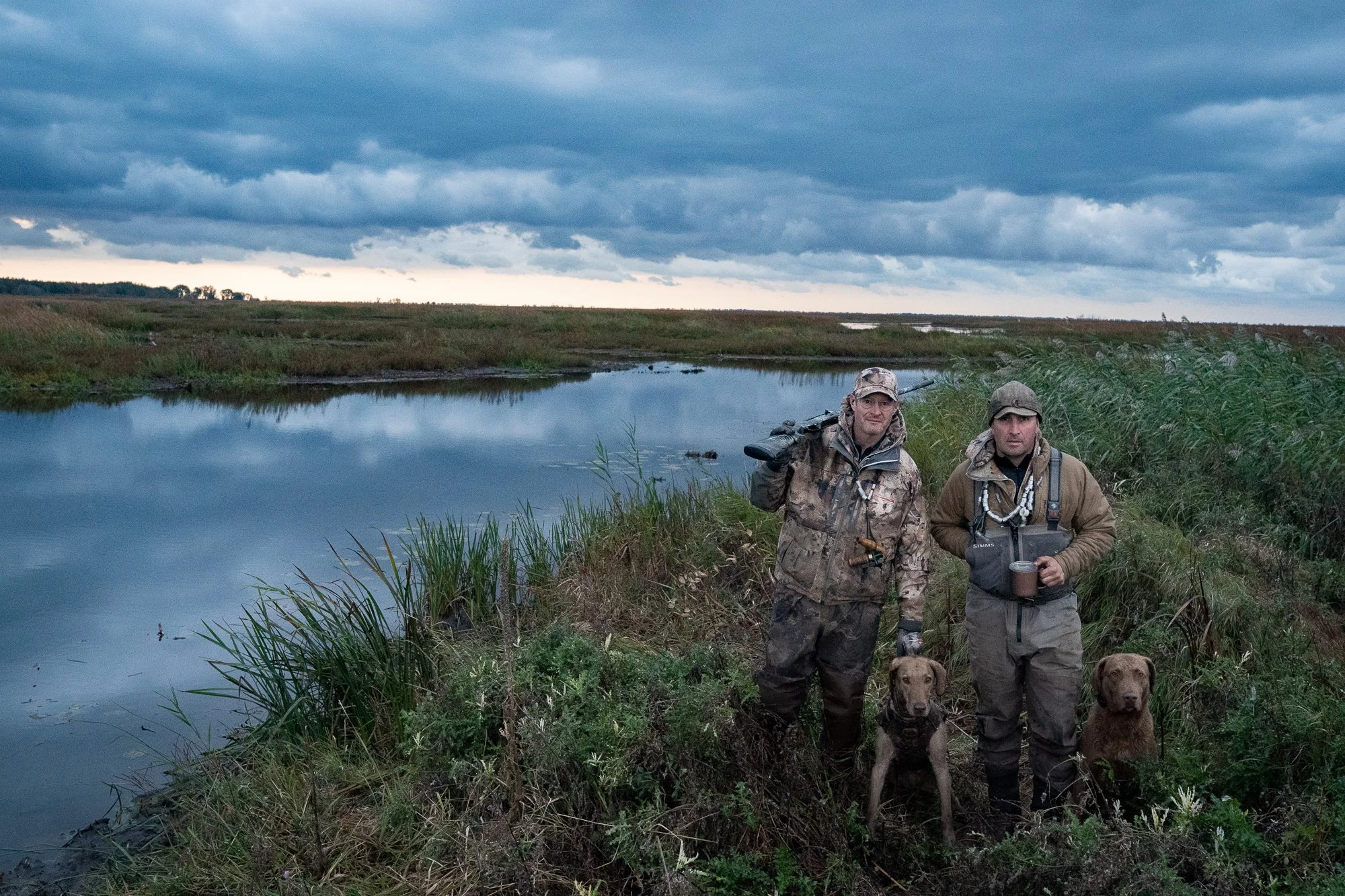 Duck hunters and retrievers stand beside marsh water after a day of waterfowl hunting in Ontario