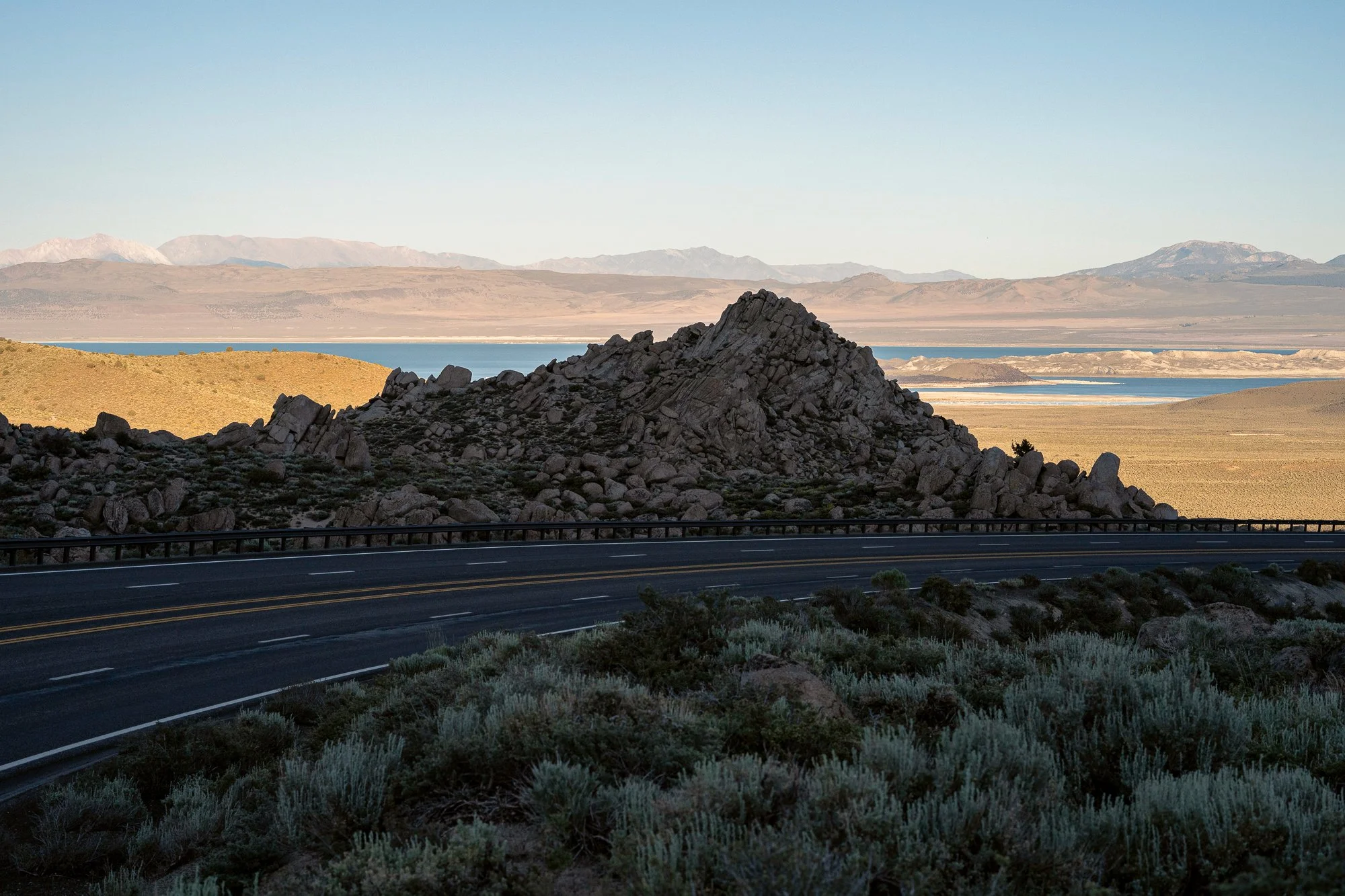 California desert highway with rocky hills, distant mountains, and blue water beyond the road