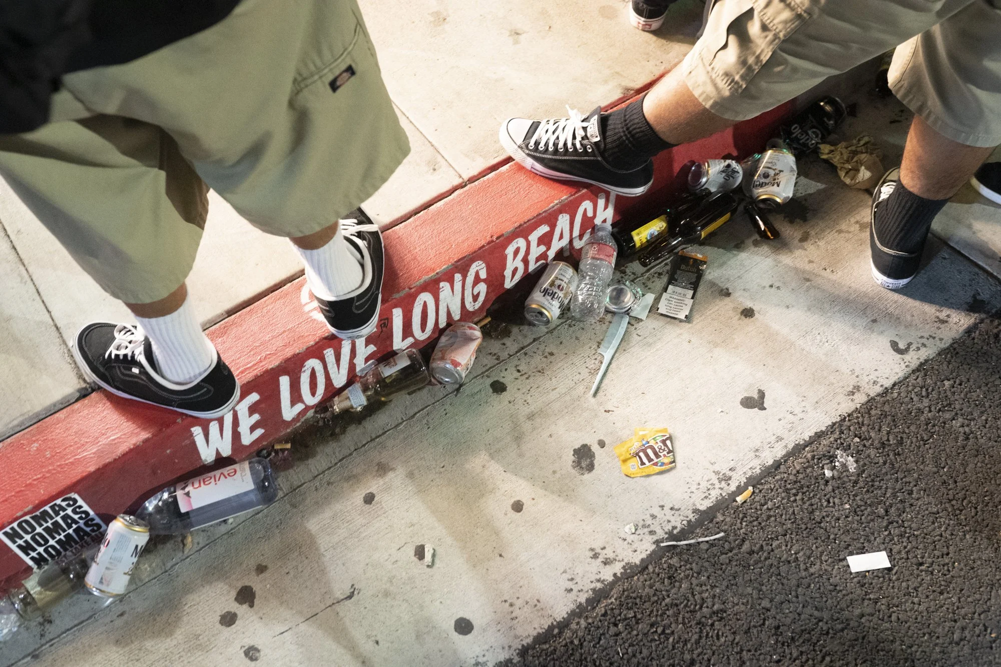 Painted curb reading “We Love Long Beach” with bottles and litter scattered along a sidewalk after a street gathering