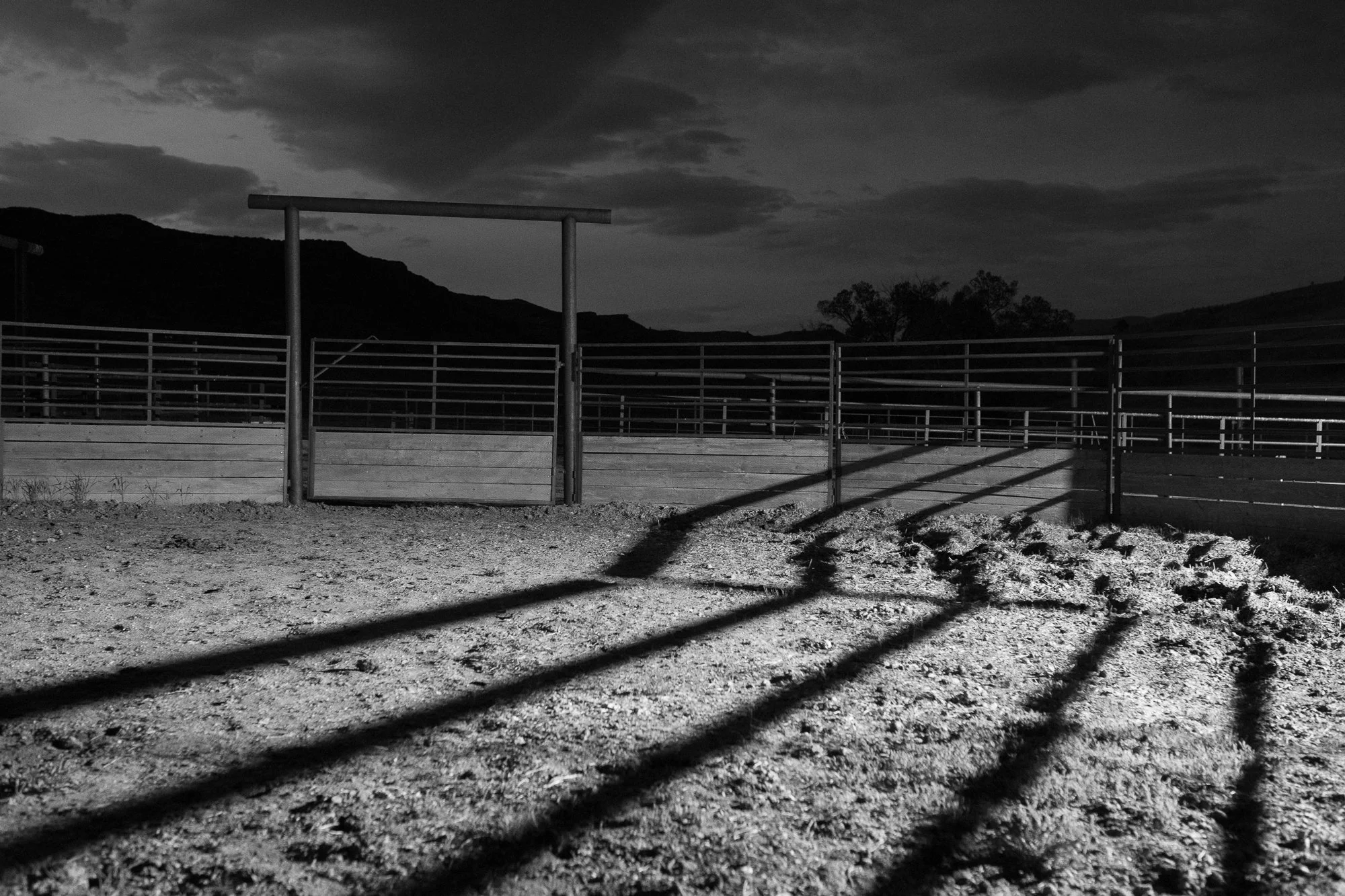 Black and white photograph of an empty cattle corral with long shadows cast across the ground
