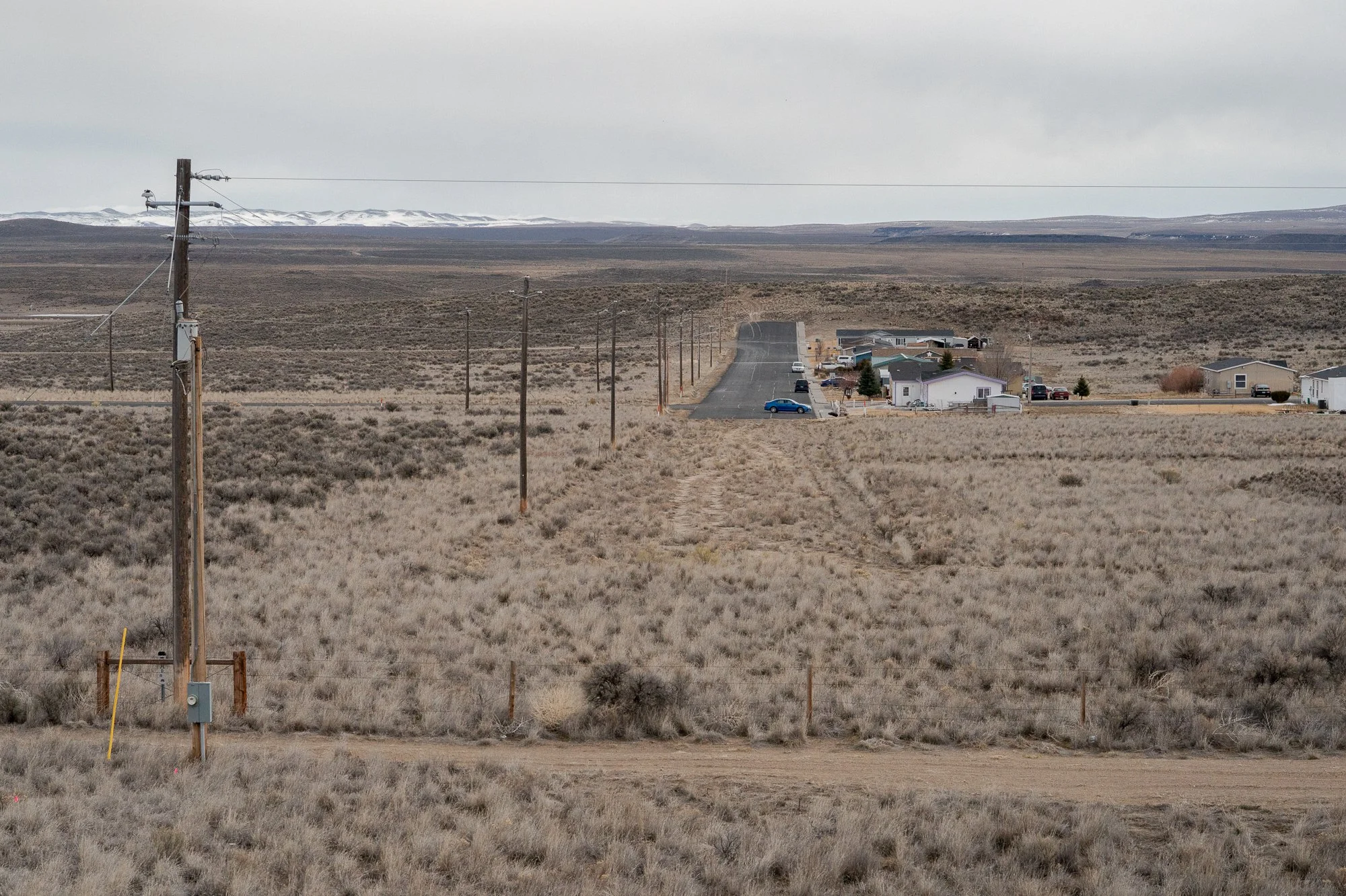 Remote Nevada small town viewed across open sagebrush flats, with a utility pole in the foreground, a road leading into a handful of houses, and snow-covered mountains on the horizon.