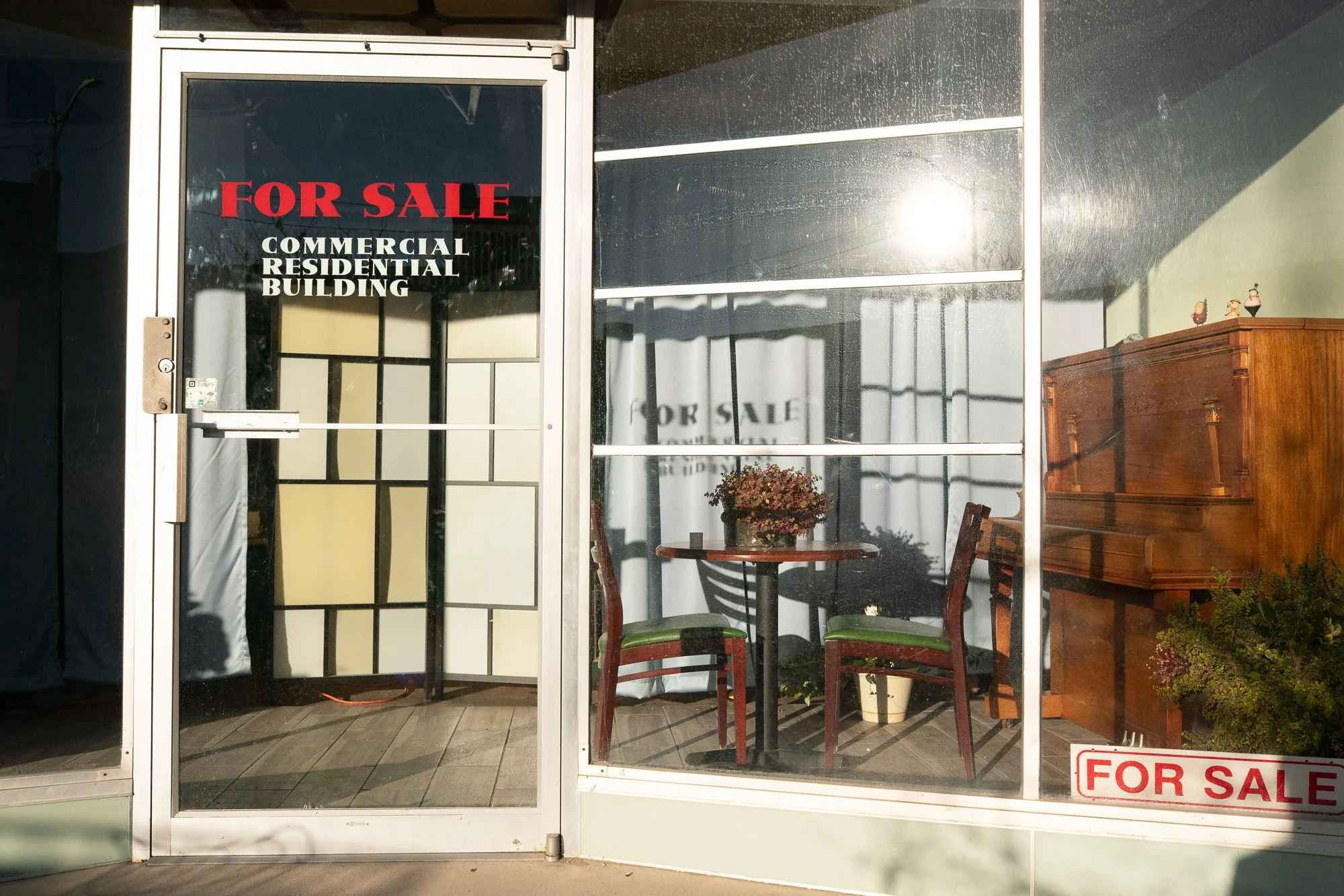 Piano, chairs, and a table visible through the windows of a commercial storefront with several for sale signs.