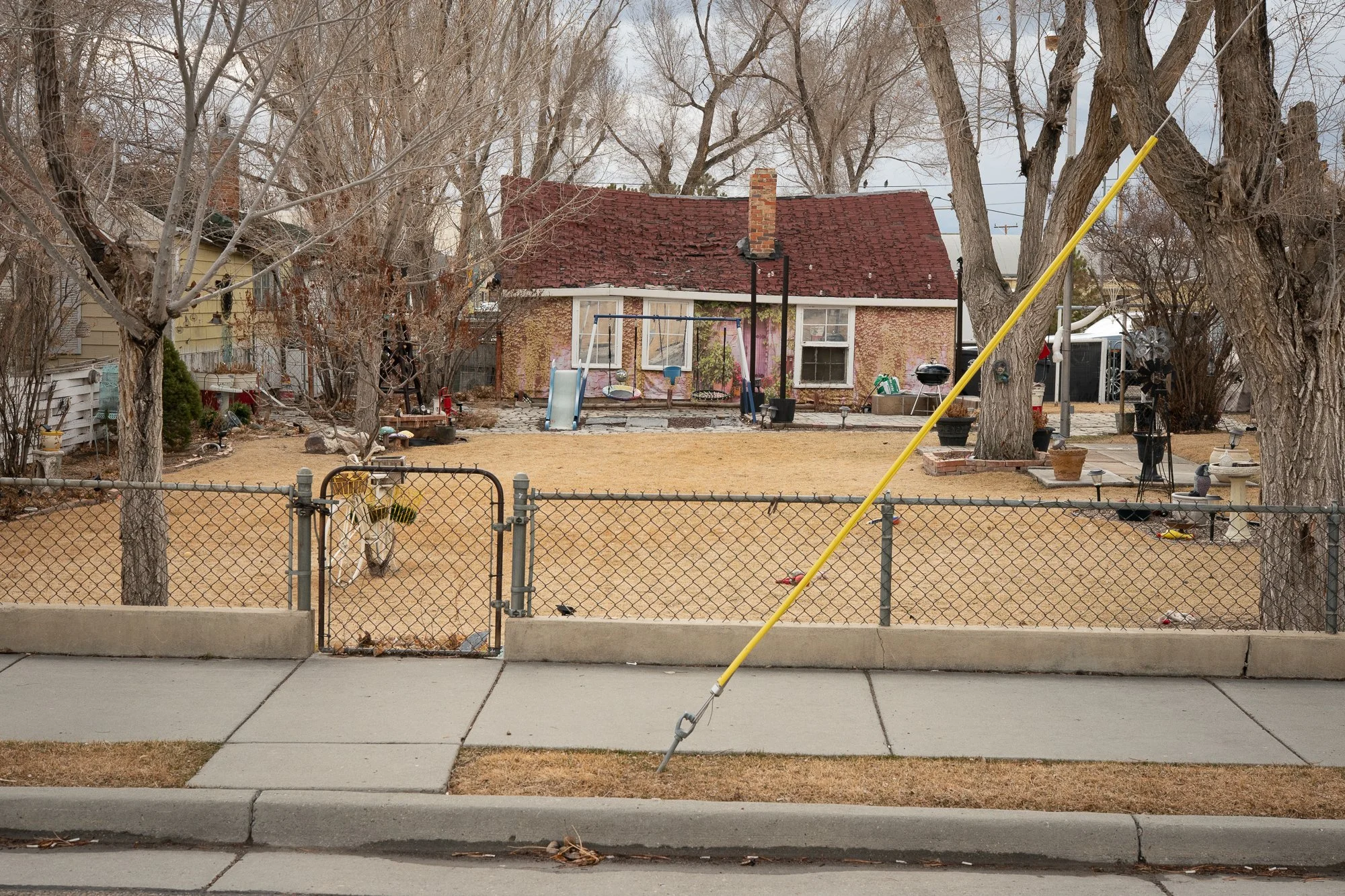 Red-roofed brick house with a fenced front yard, leafless trees, and play equipment beside the sidewalk.