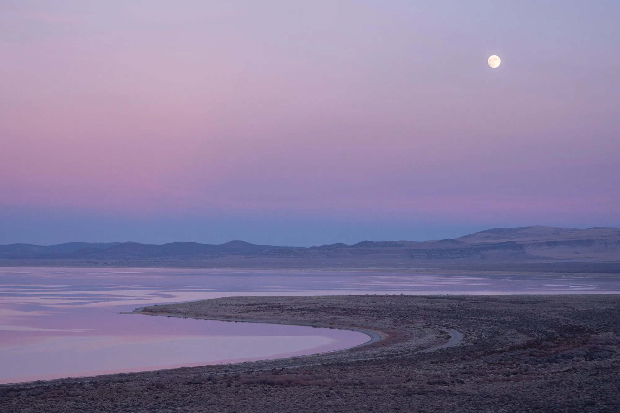 Moon above a quiet California desert lake with pink evening light and distant hills
