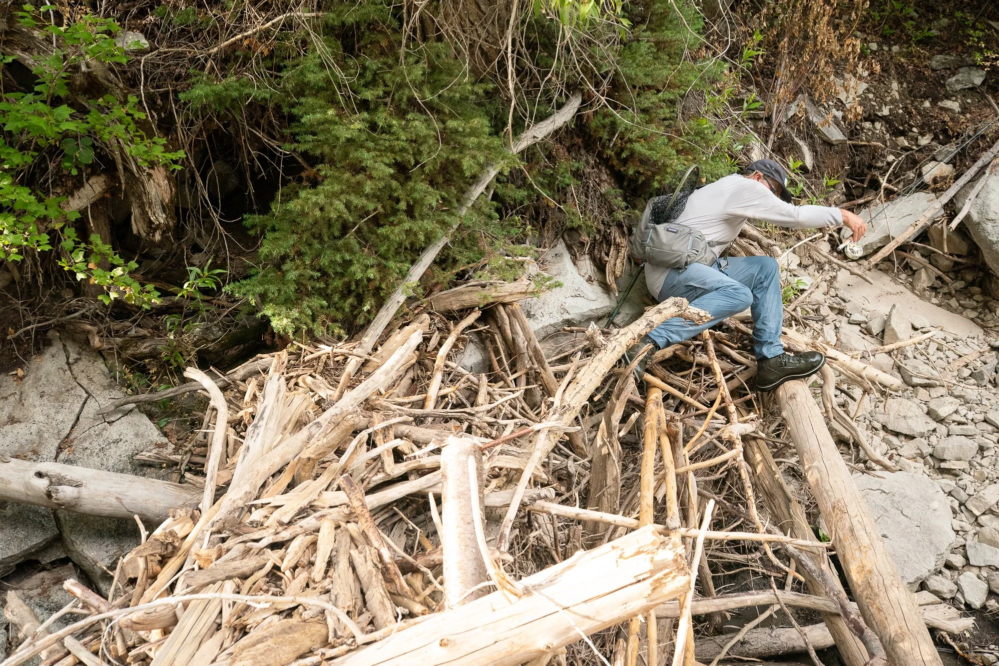 A fly angler steps across driftwood and rocks near the riverbank with a rod in hand