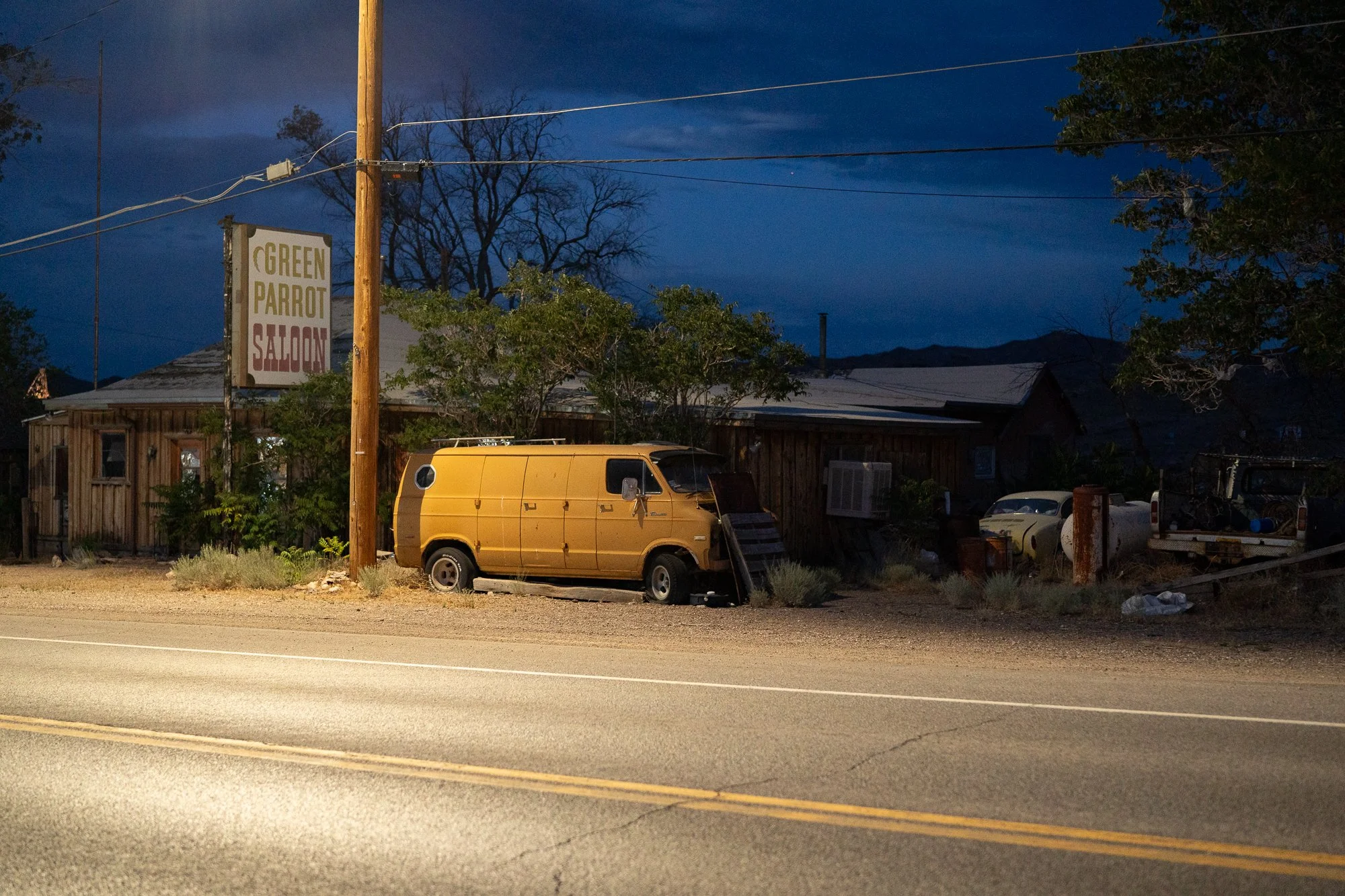 Old yellow van parked in front of the Green Parrot Saloon along a Nevada highway at night, with a weathered wood building and deep blue sky above the trees.