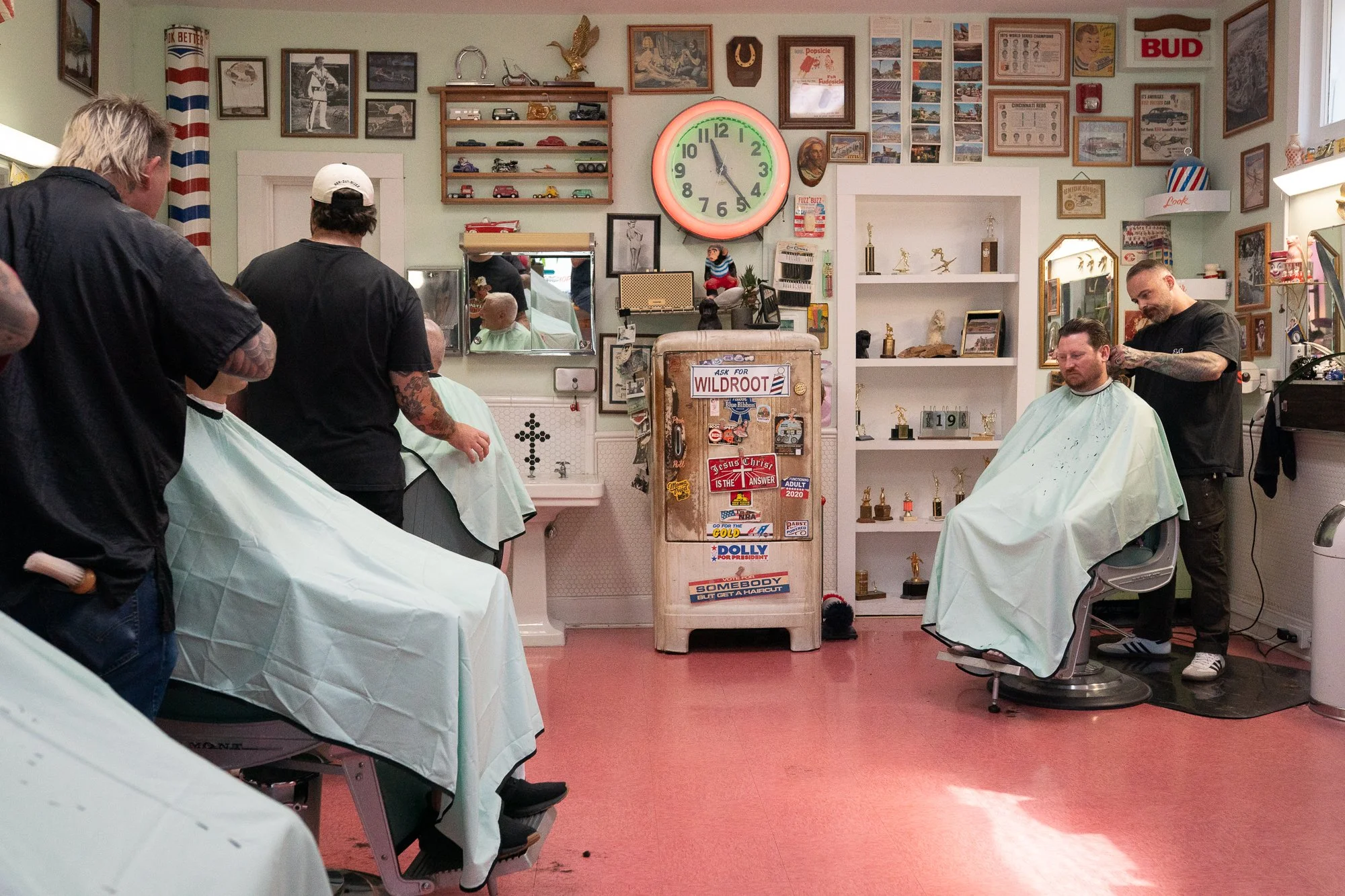 Wide interior view of Spanky’s Barbershop in Covington, Kentucky featuring vintage neon clock, Wildroot refrigerator, and framed wall art.