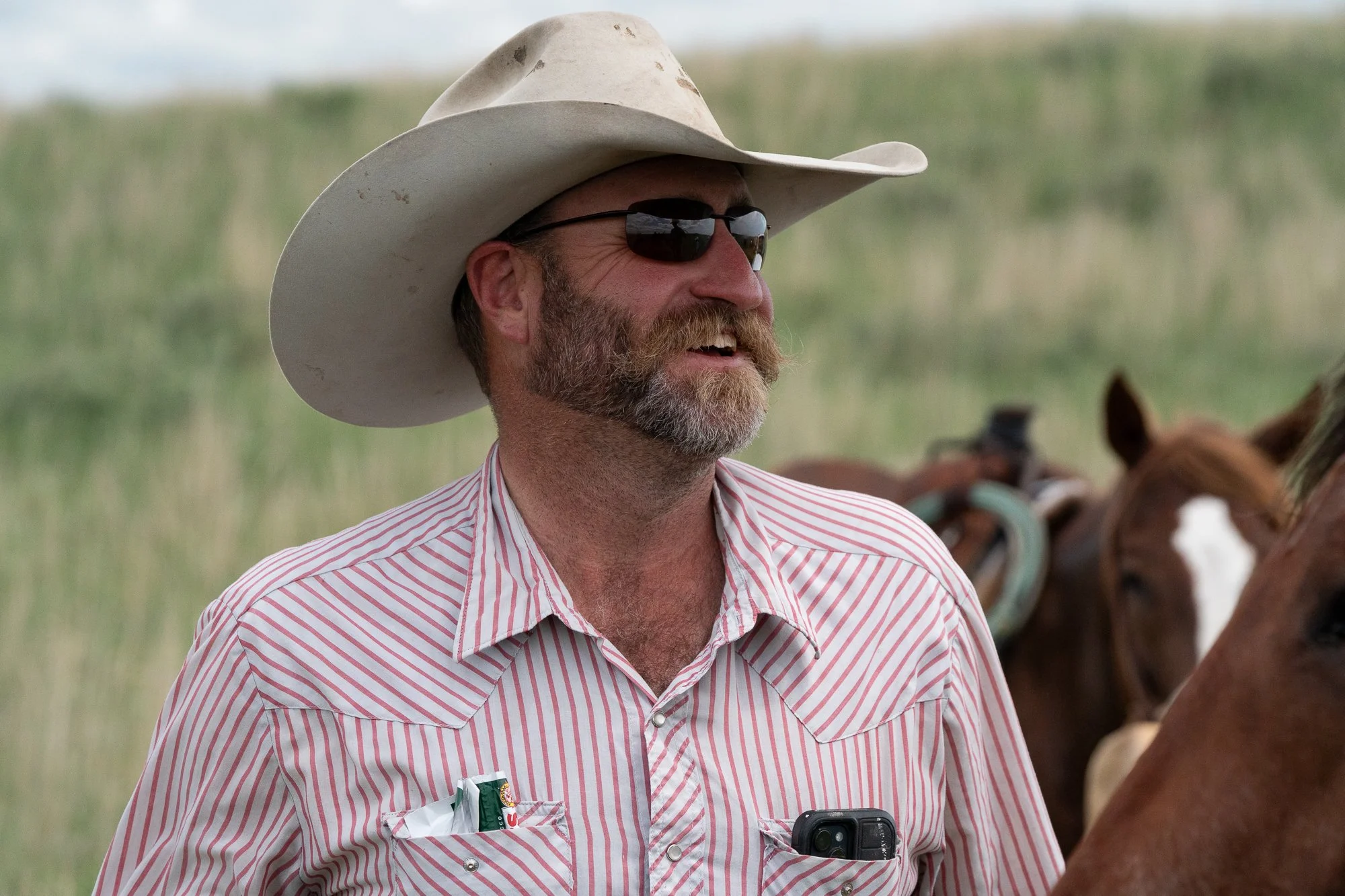 Cowboy wearing a white hat and sunglasses laughing while standing beside saddled horses at Haythorn Ranch in the Nebraska Sandhills.
