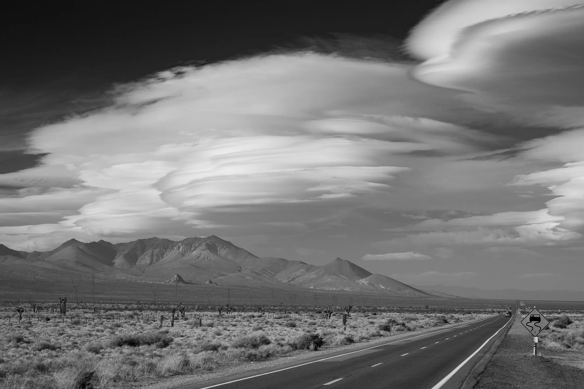Black and white California desert highway with mountains, clouds, and roadside sign