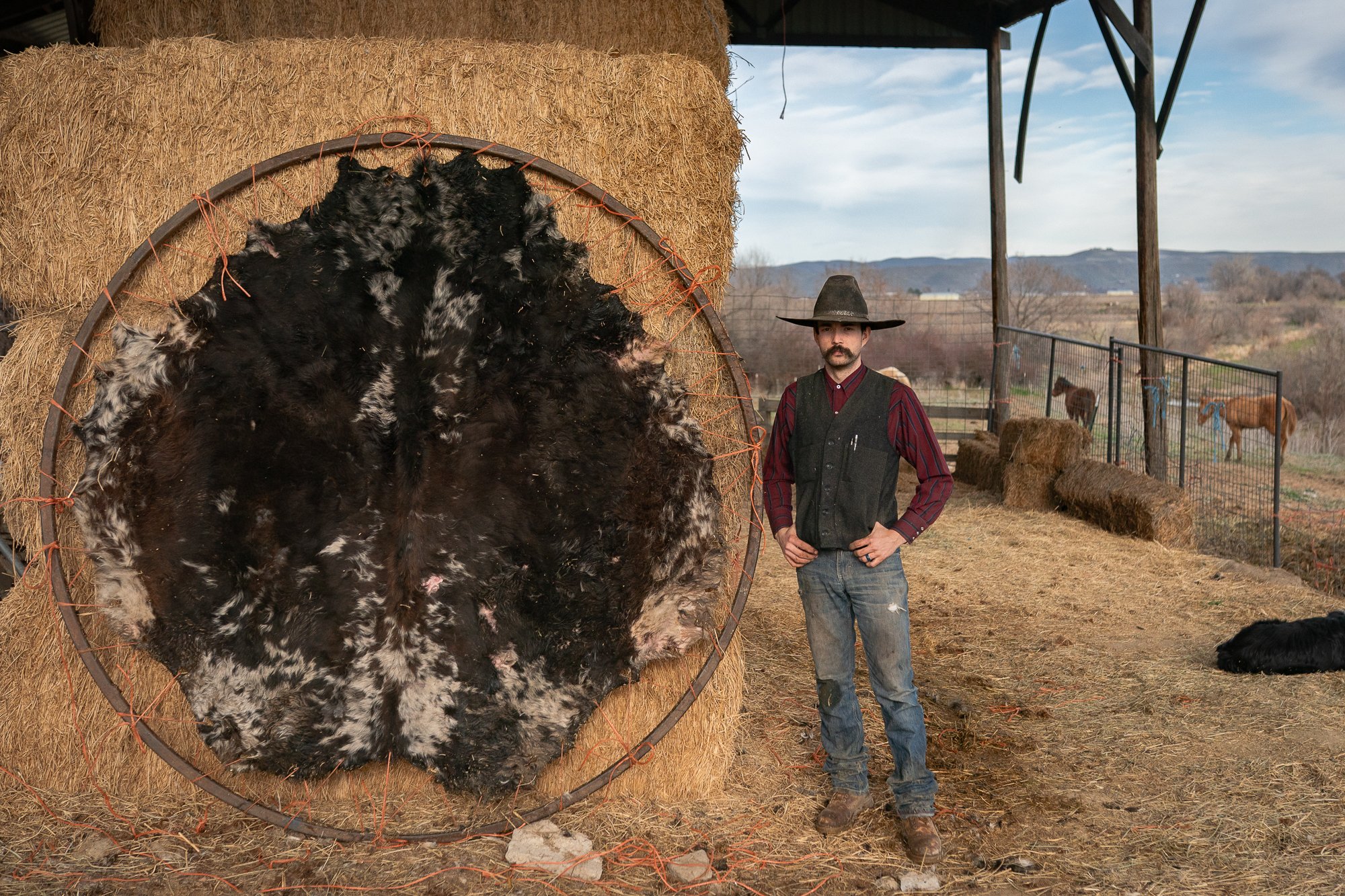 Dakota May standing beside a stretched rawhide hide drying on a round frame outside his workshop