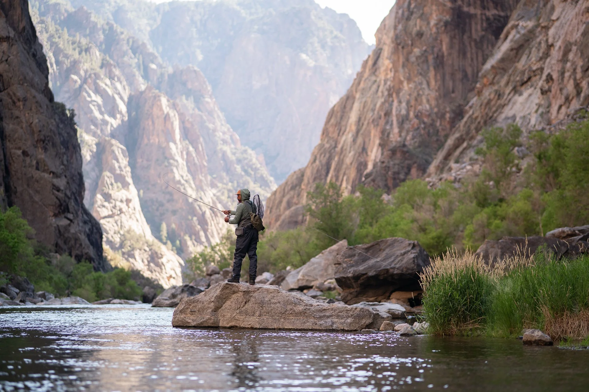 A fly angler casts from a rock in a canyon river with tall cliffs in the background