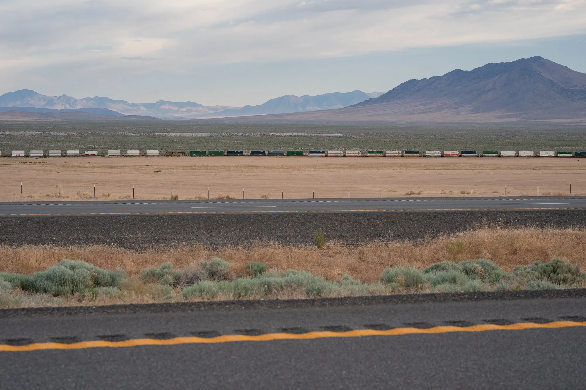 Long freight train running parallel to a Nevada highway across a wide desert basin, with sagebrush in the foreground and multiple mountain ranges visible in the distance.