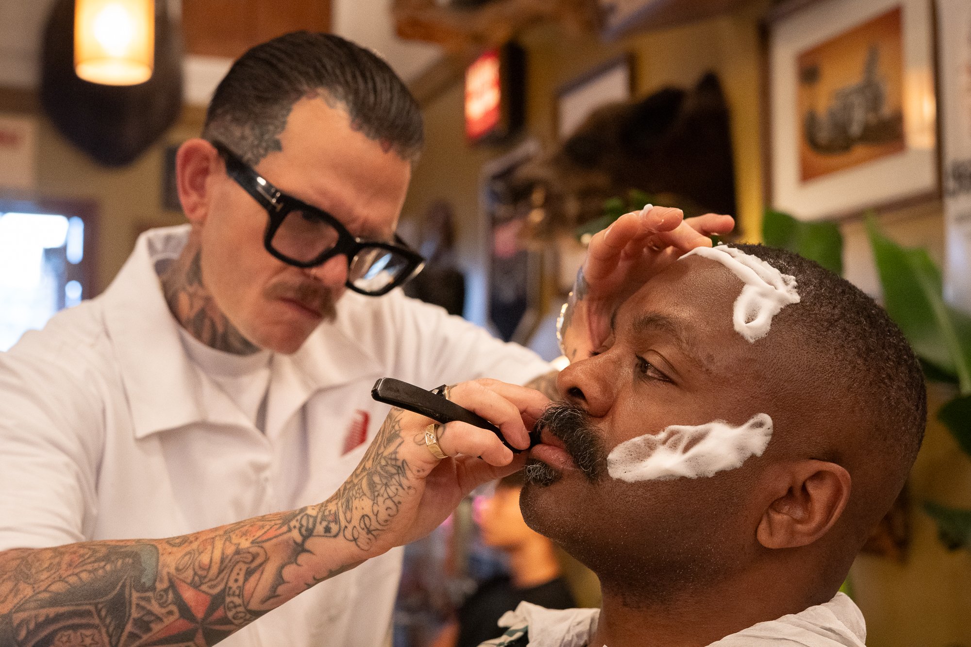A barber in a white uniform uses a straight razor to shave a client's beard at Pugsly's barbershop.