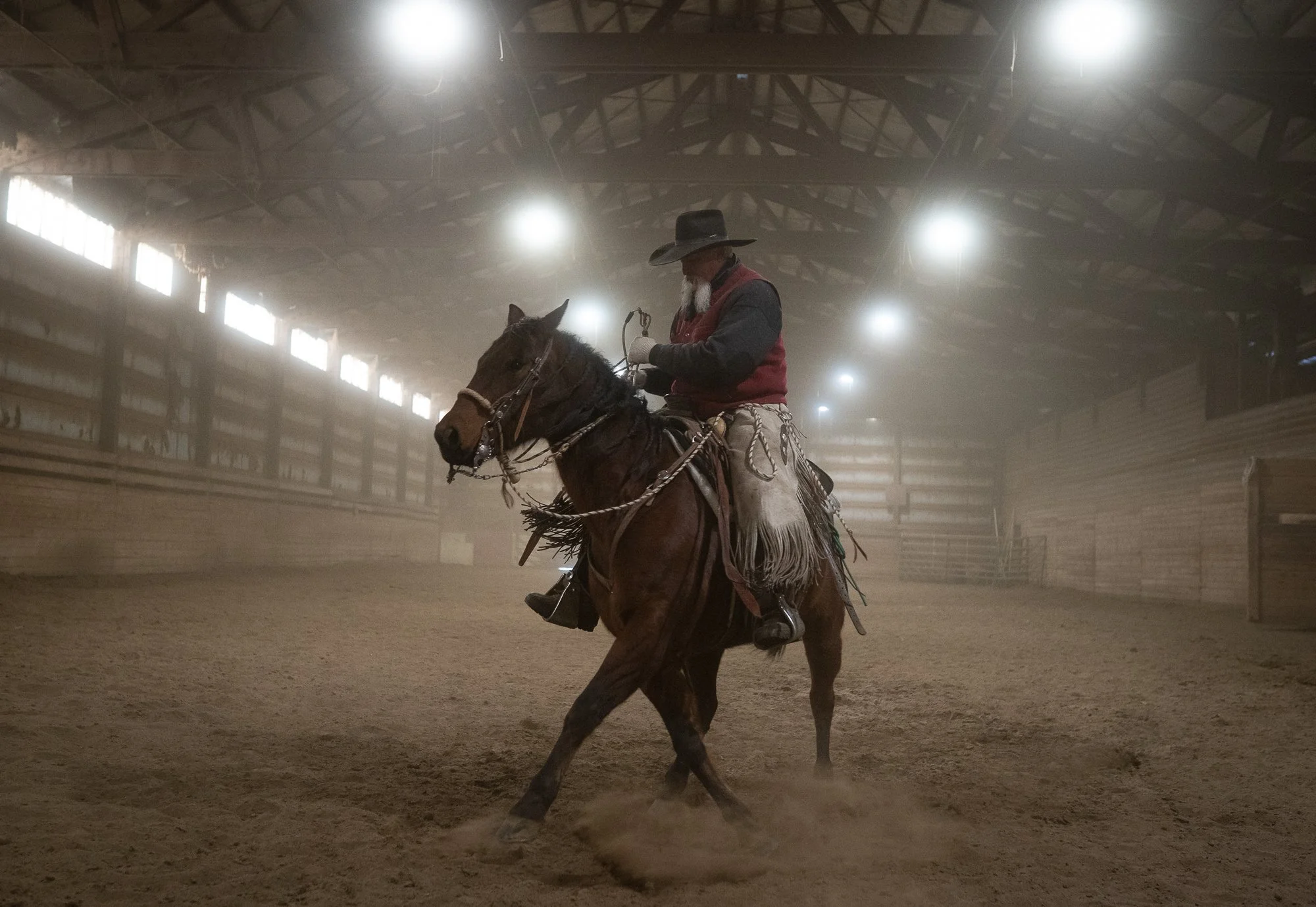 Cowboy riding horse inside dusty indoor arena with dramatic overhead lights