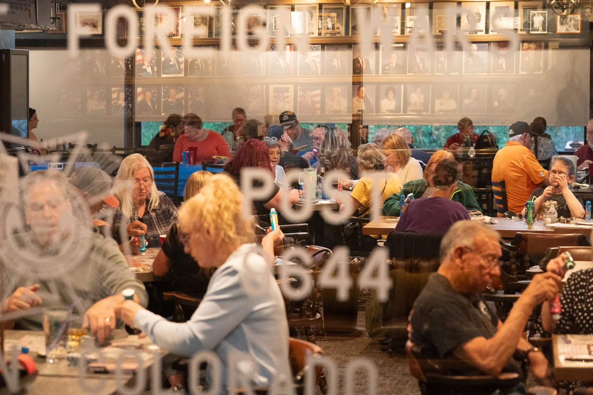 People gathered inside a VFW hall seen through the front window with portraits on the wall