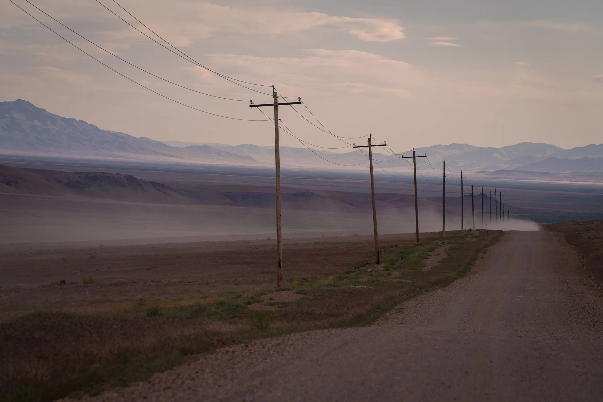 Straight Nevada dirt road lined with wooden utility poles receding into a hazy desert basin, with blowing dust and distant mountain ranges under a pale sky.