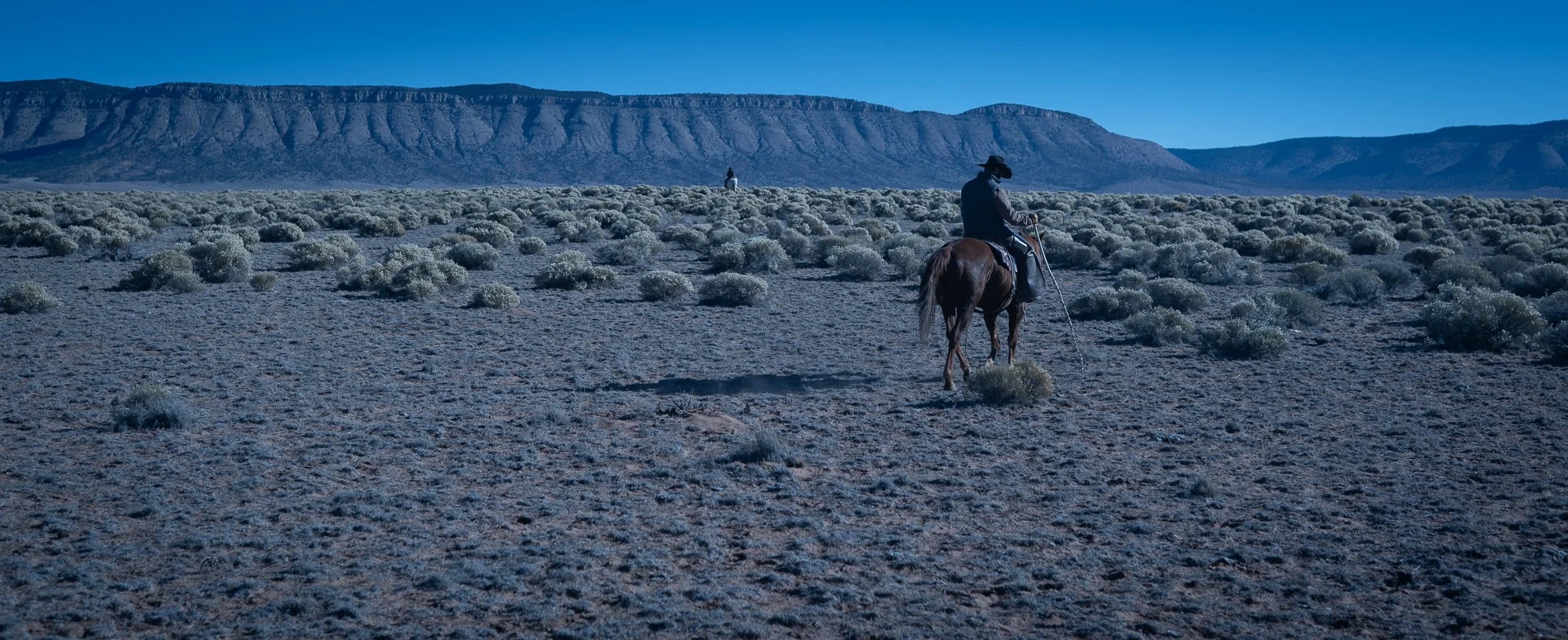 Photograph of two cowboys riding their horses through a vast desert landscape at night