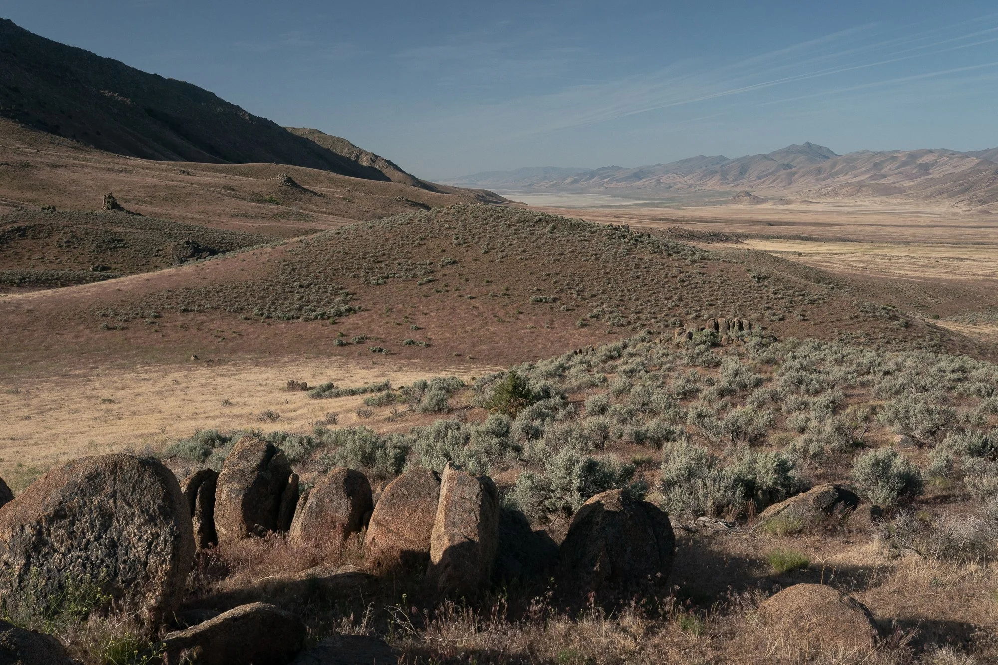 Rocky Nevada hillside overlooking a wide desert valley with sagebrush, boulders, and distant mountain ranges under a clear blue sky.
