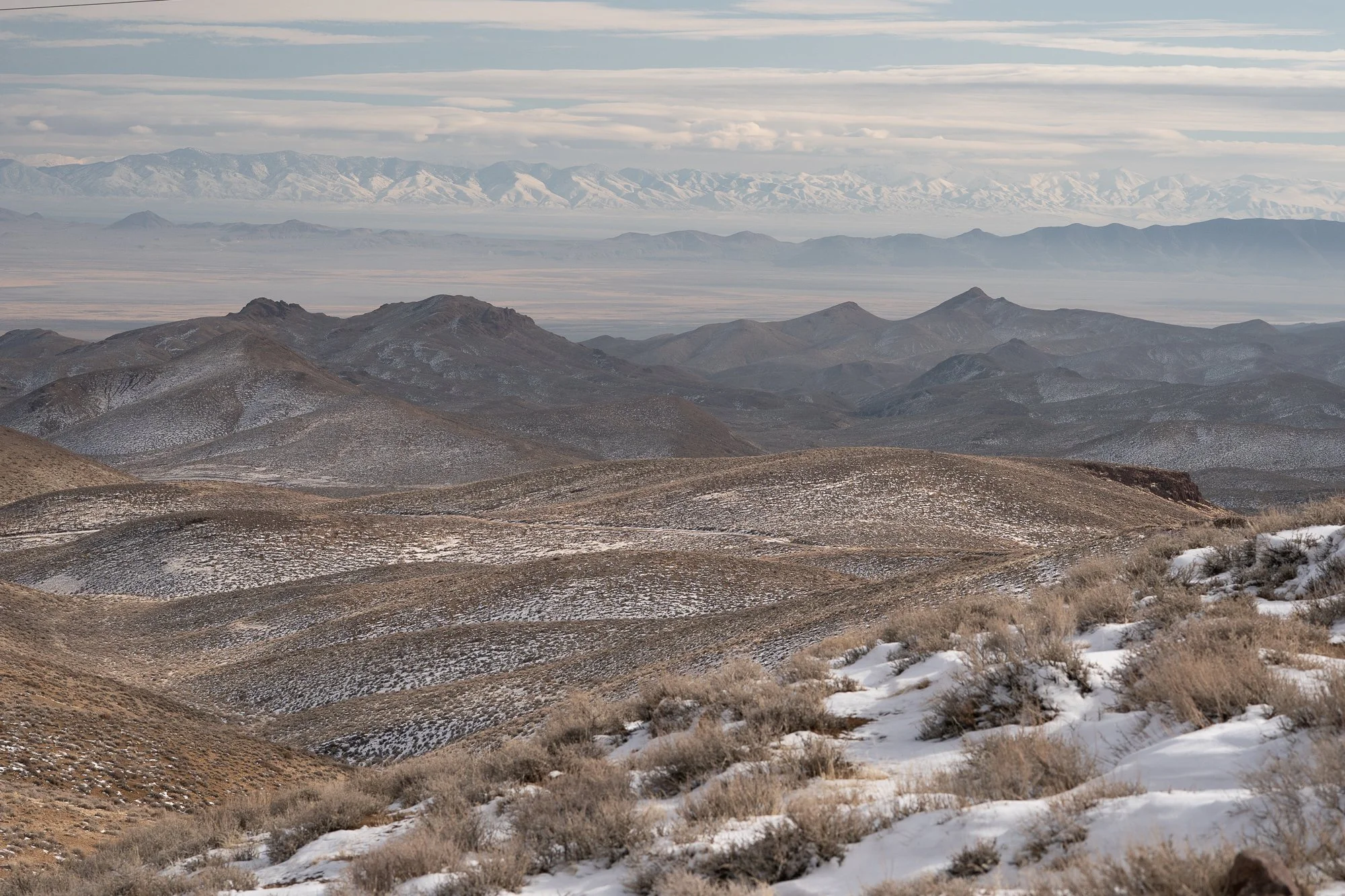 Wide view of multiple Nevada mountain ranges with light snow cover on the ridges, dry scrub in the foreground, and distant peaks fading into atmospheric haze.