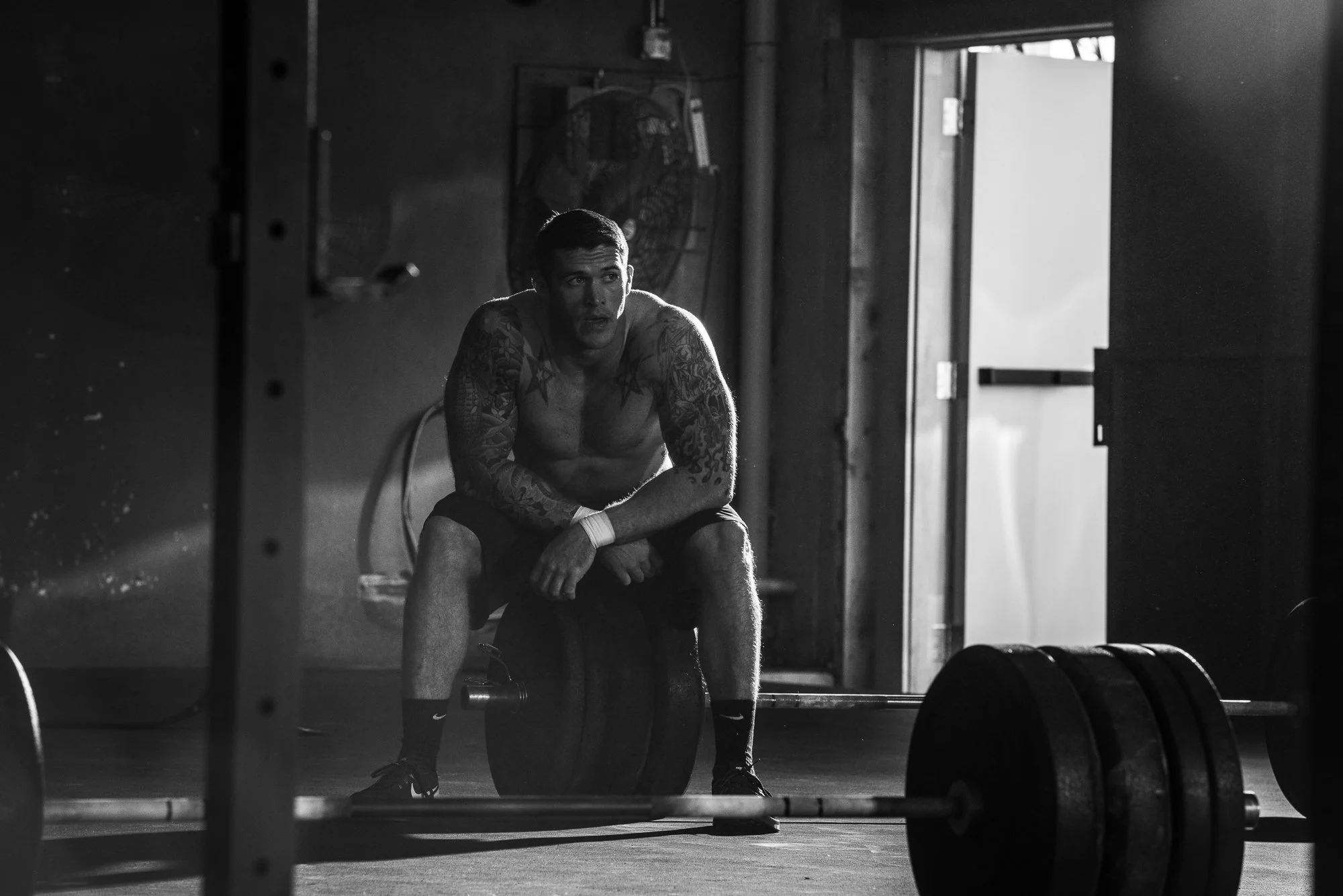CrossFit athlete resting on a wooden plyometric box beside loaded barbells in a gritty gym.