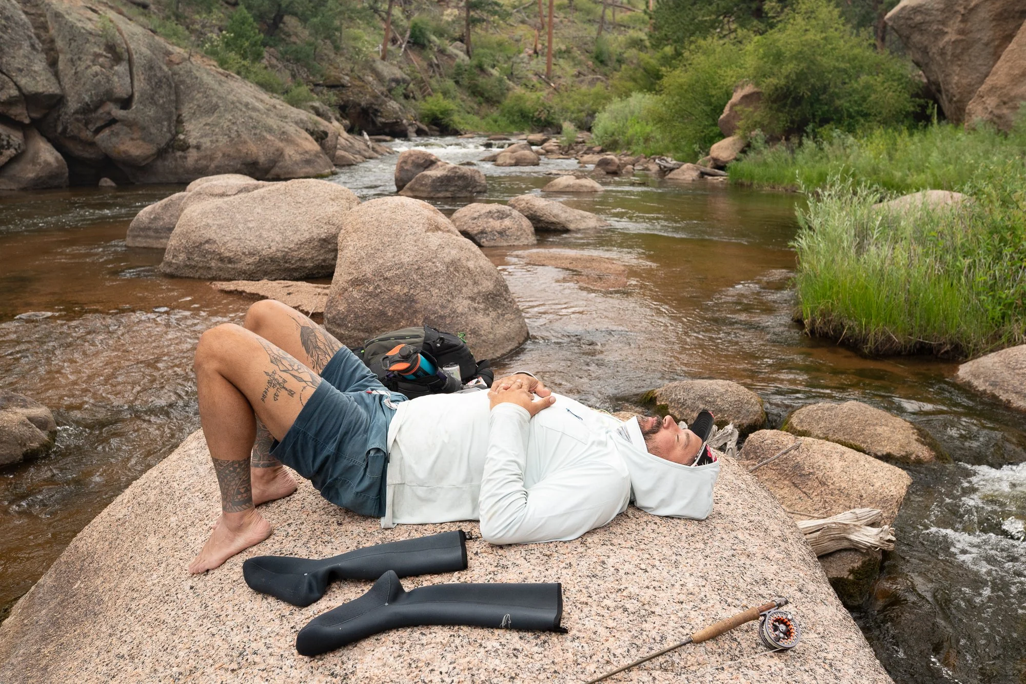 A fly angler rests on a large rock beside the river with his rod and gear nearby