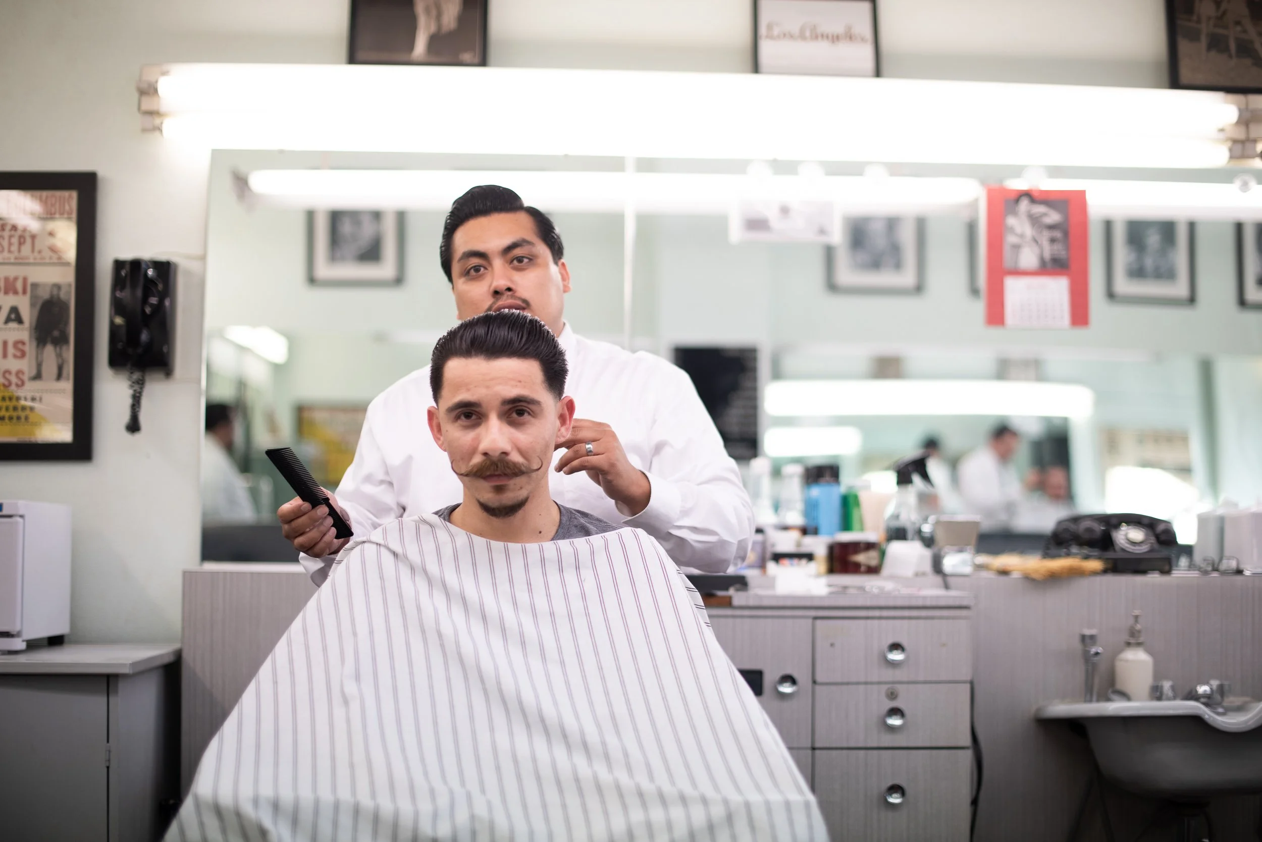 Barber in white shirt standing behind a caped client with a slicked pompadour and waxed mustache at Sweeney Todd's Barbershop, both facing the camera