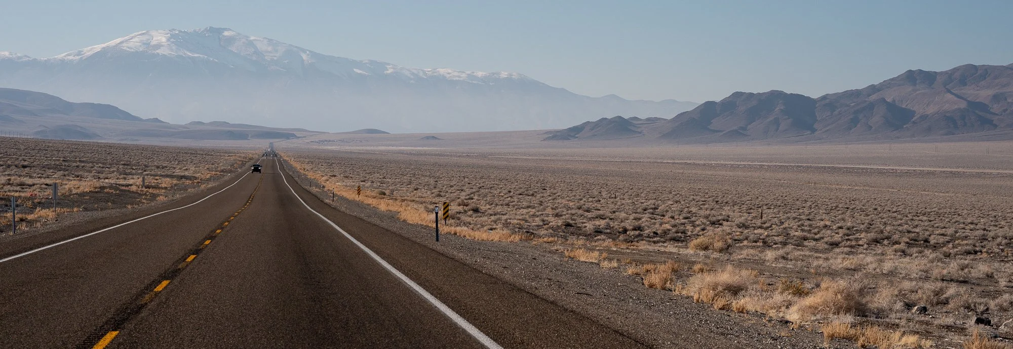 Wide Nevada highway stretching toward the horizon with one car in the distance, a large snow-capped mountain to the left, and open desert basin on both sides under a pale hazy sky.