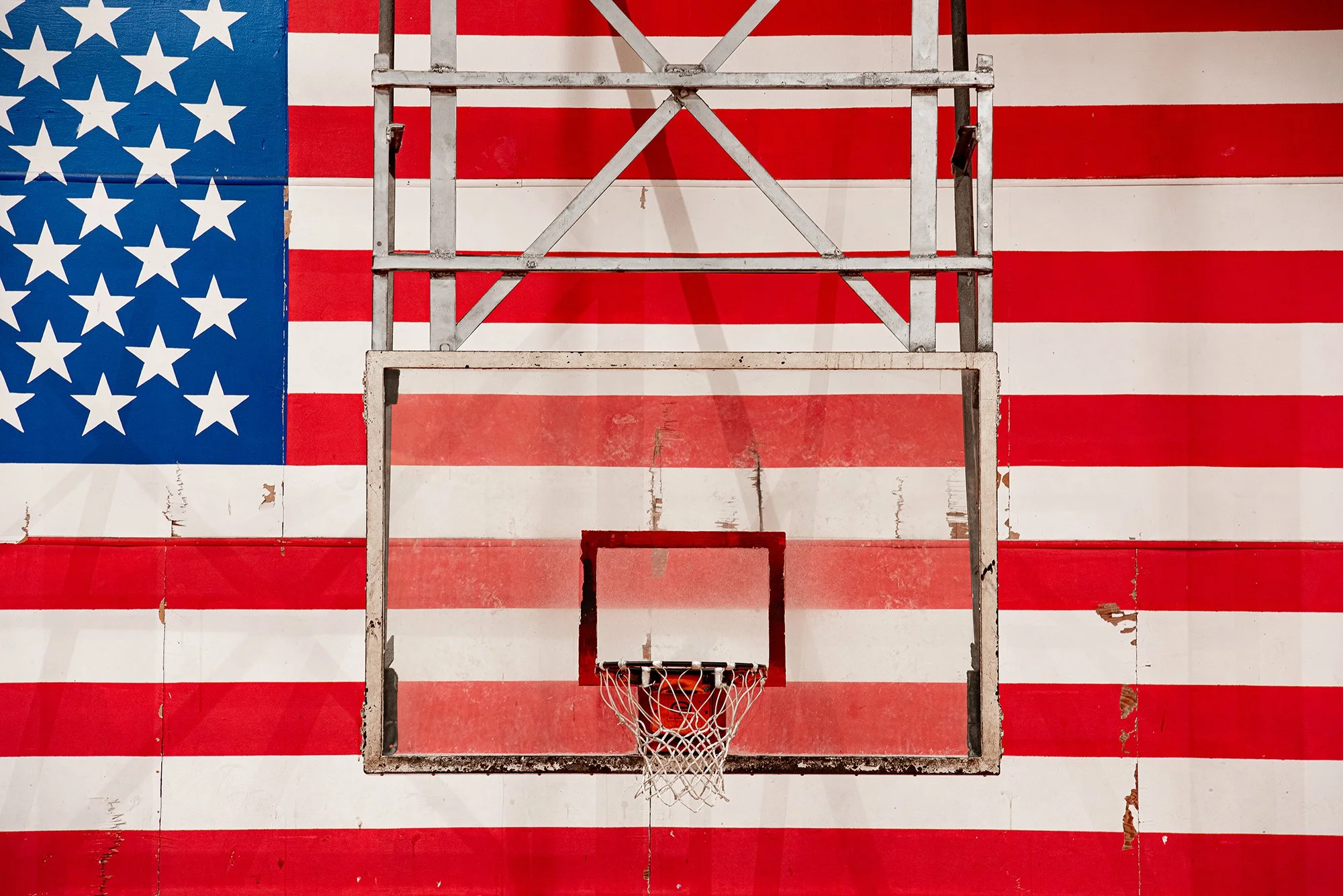 Weathered basketball backboard and hoop mounted against a large American flag mural.