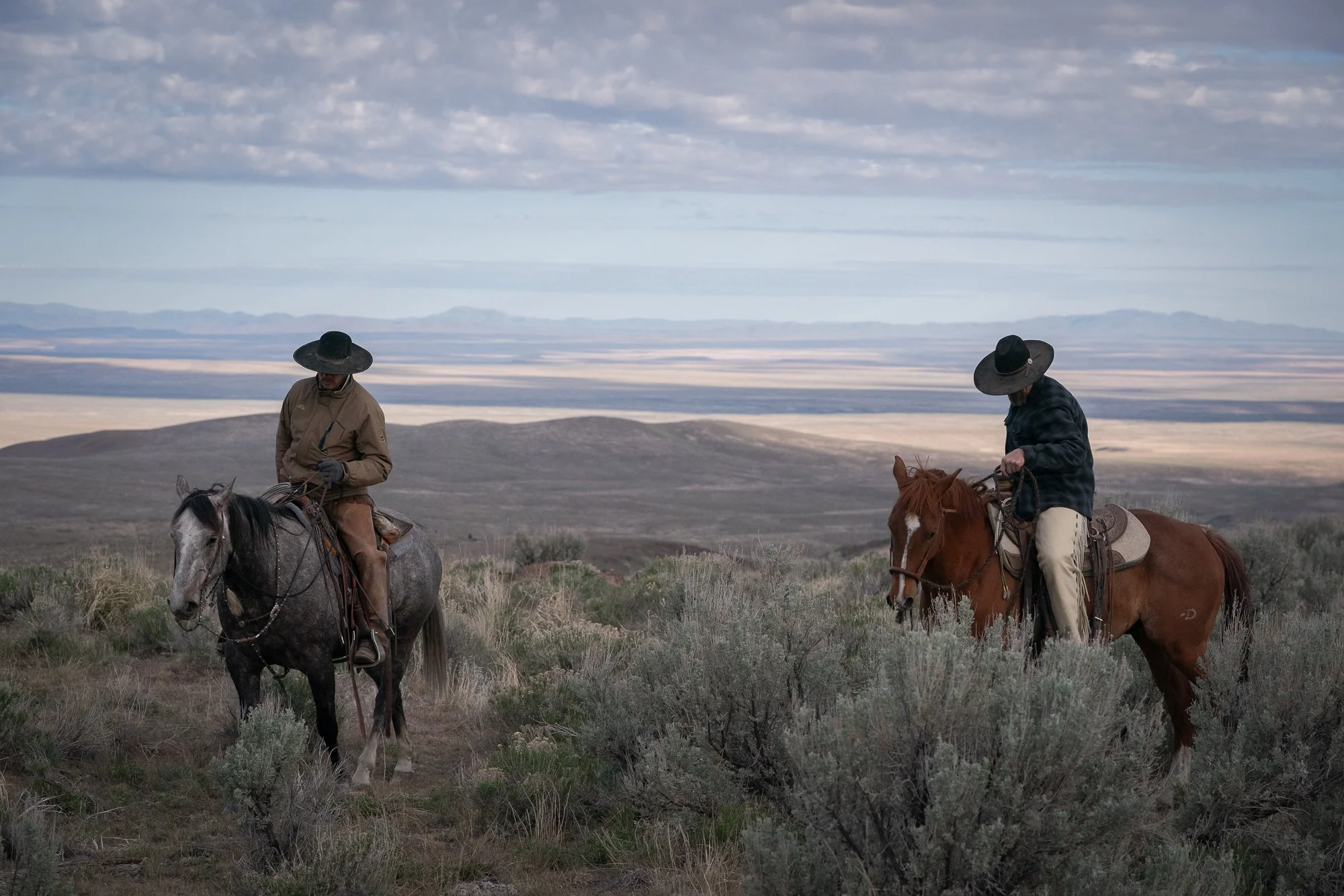 Two buckaroos on horseback riding through sagebrush across the open Great Basin landscape.