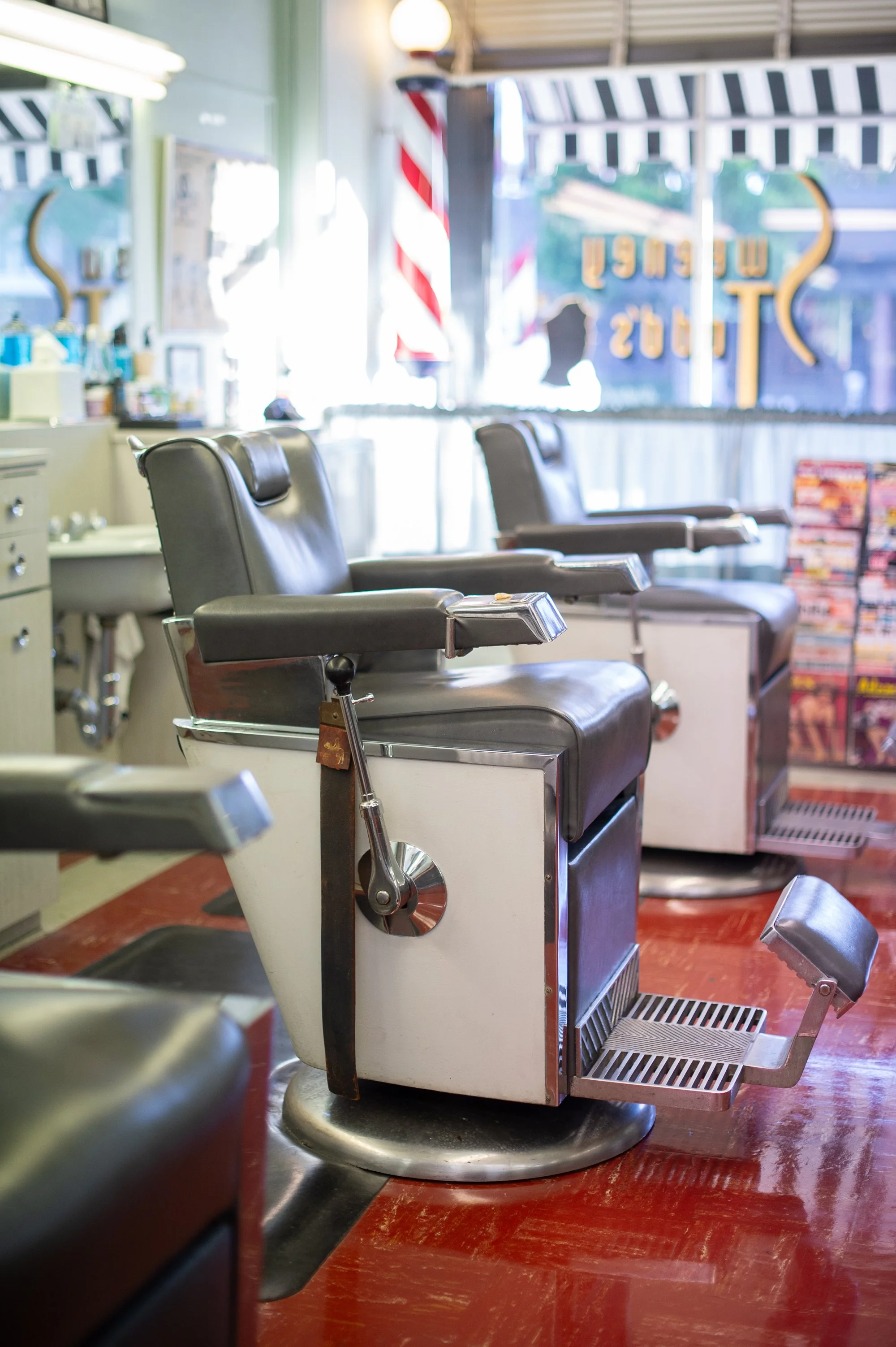 Row of empty vintage chrome and leather barber chairs at Sweeney Todd's Barbershop with barber pole and Sweeney Todd's gold window lettering reflected behind them