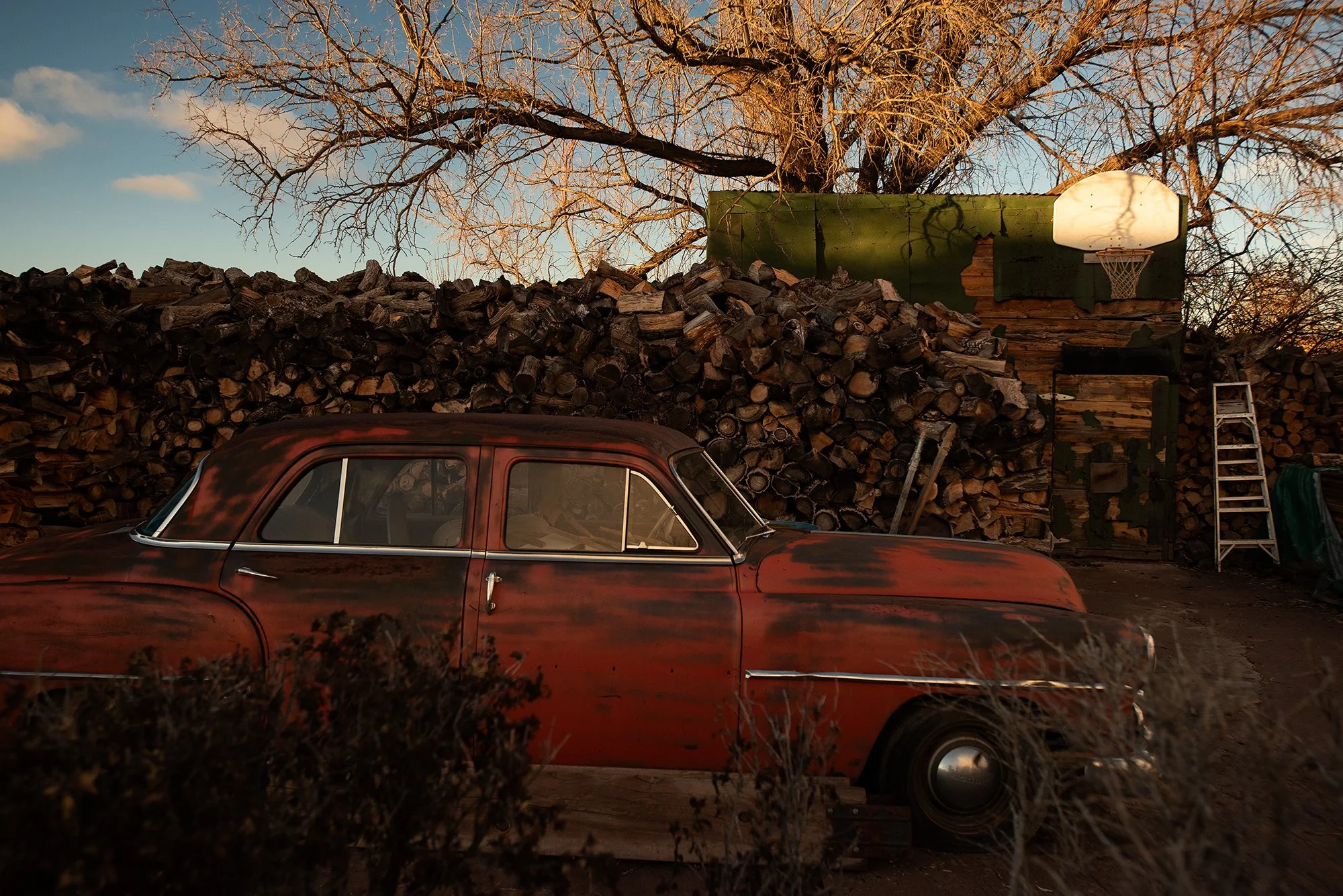 Weathered basketball hoop mounted above a woodpile beside a vintage car and a large green structure.